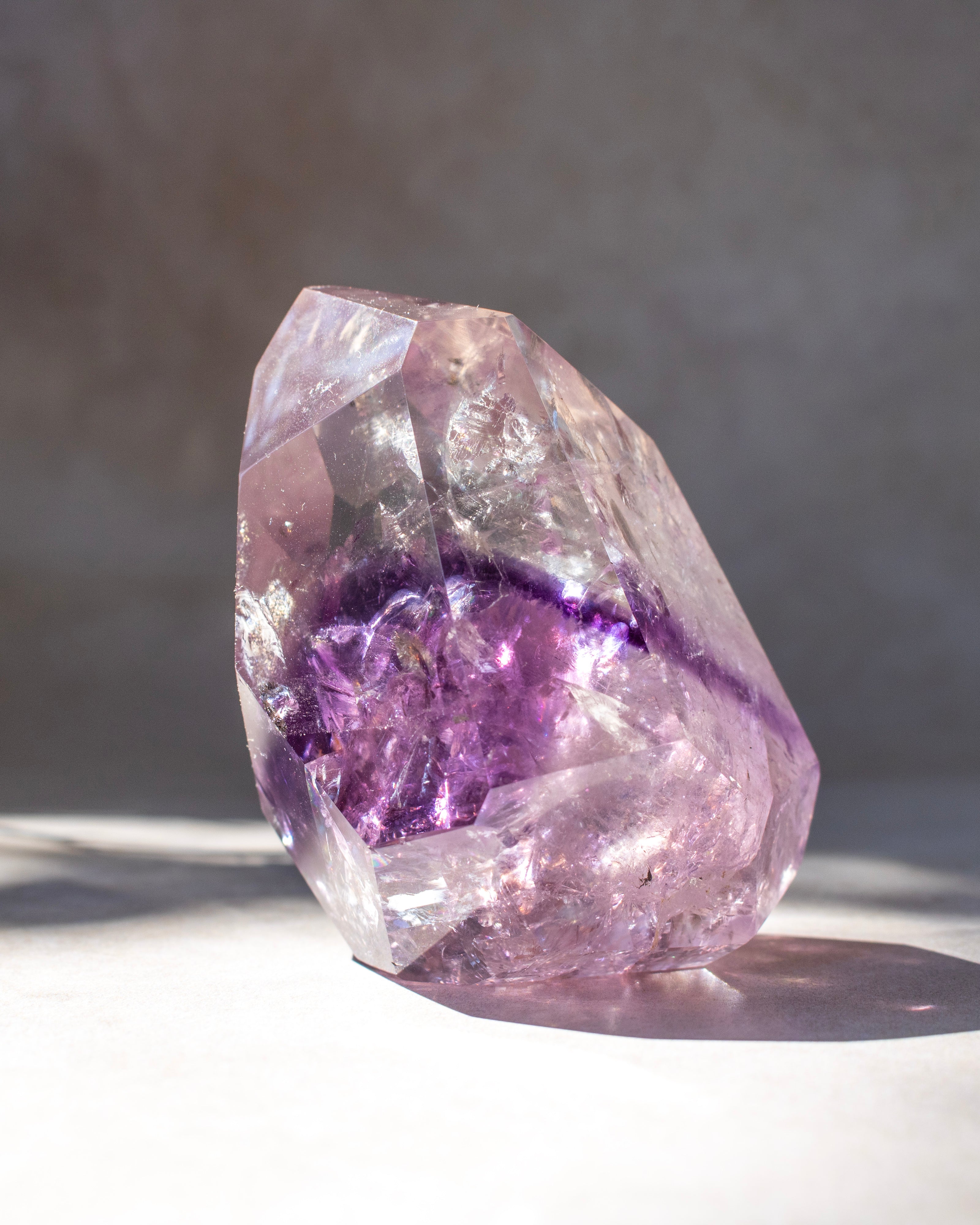 A natural amethyst crystal cluster with a prominent purple band around the middle, photographed on a light surface with a soft gray background.