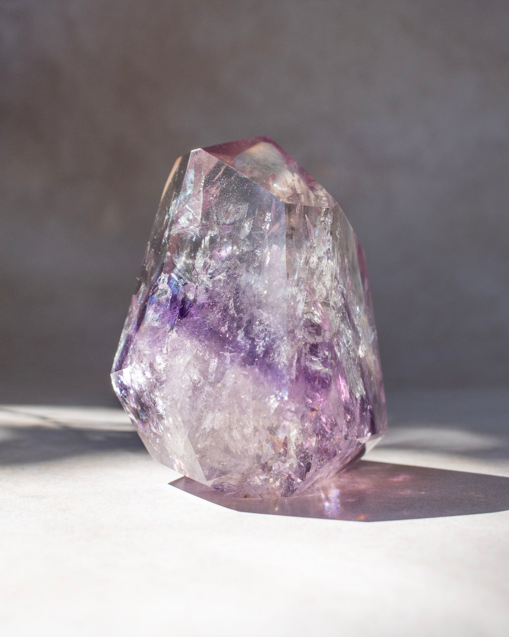 A natural amethyst crystal cluster with purple and lavender bands, standing on a light surface against a neutral gray background.