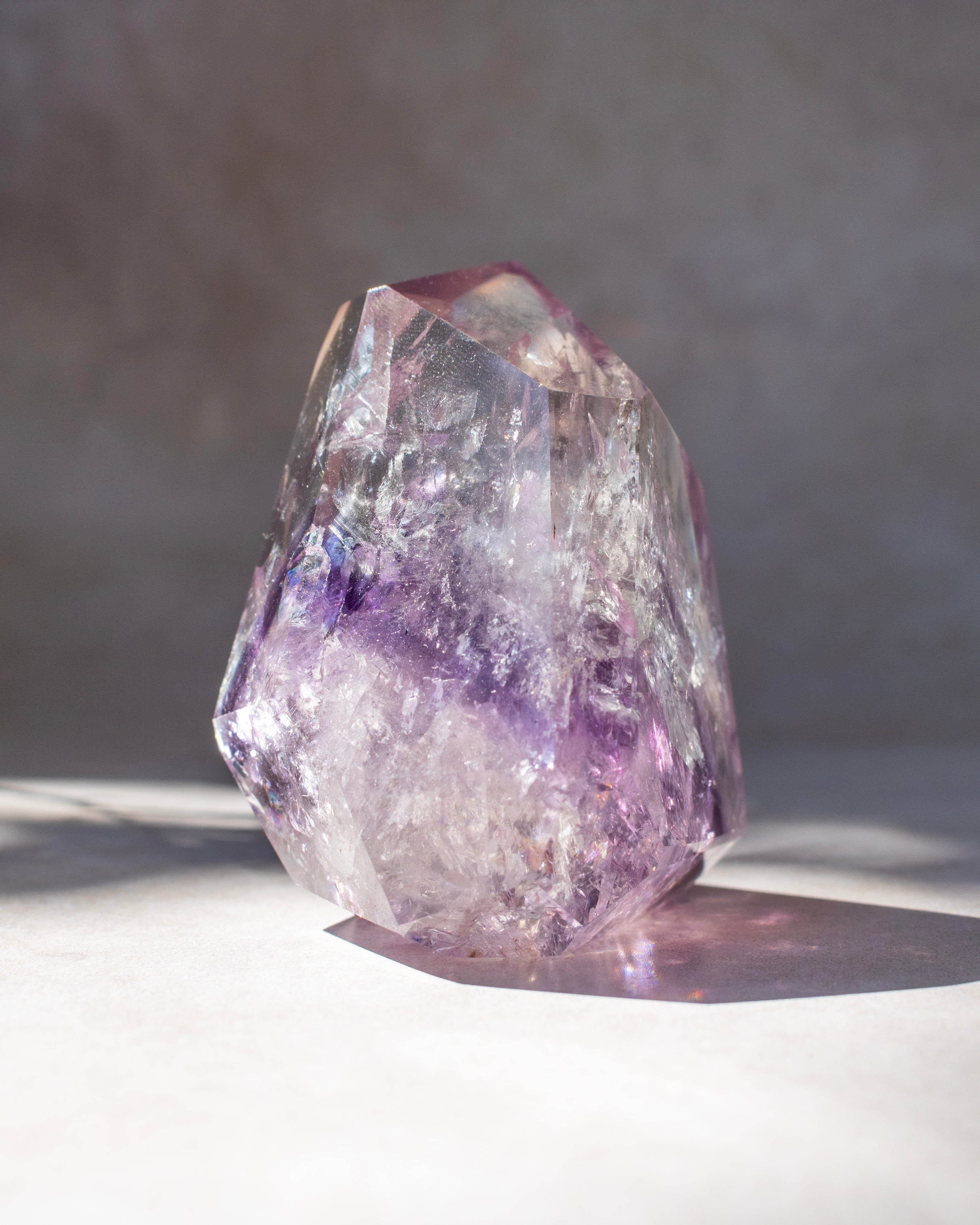 A natural amethyst crystal cluster with purple and lavender bands, standing on a light surface against a neutral gray background.
