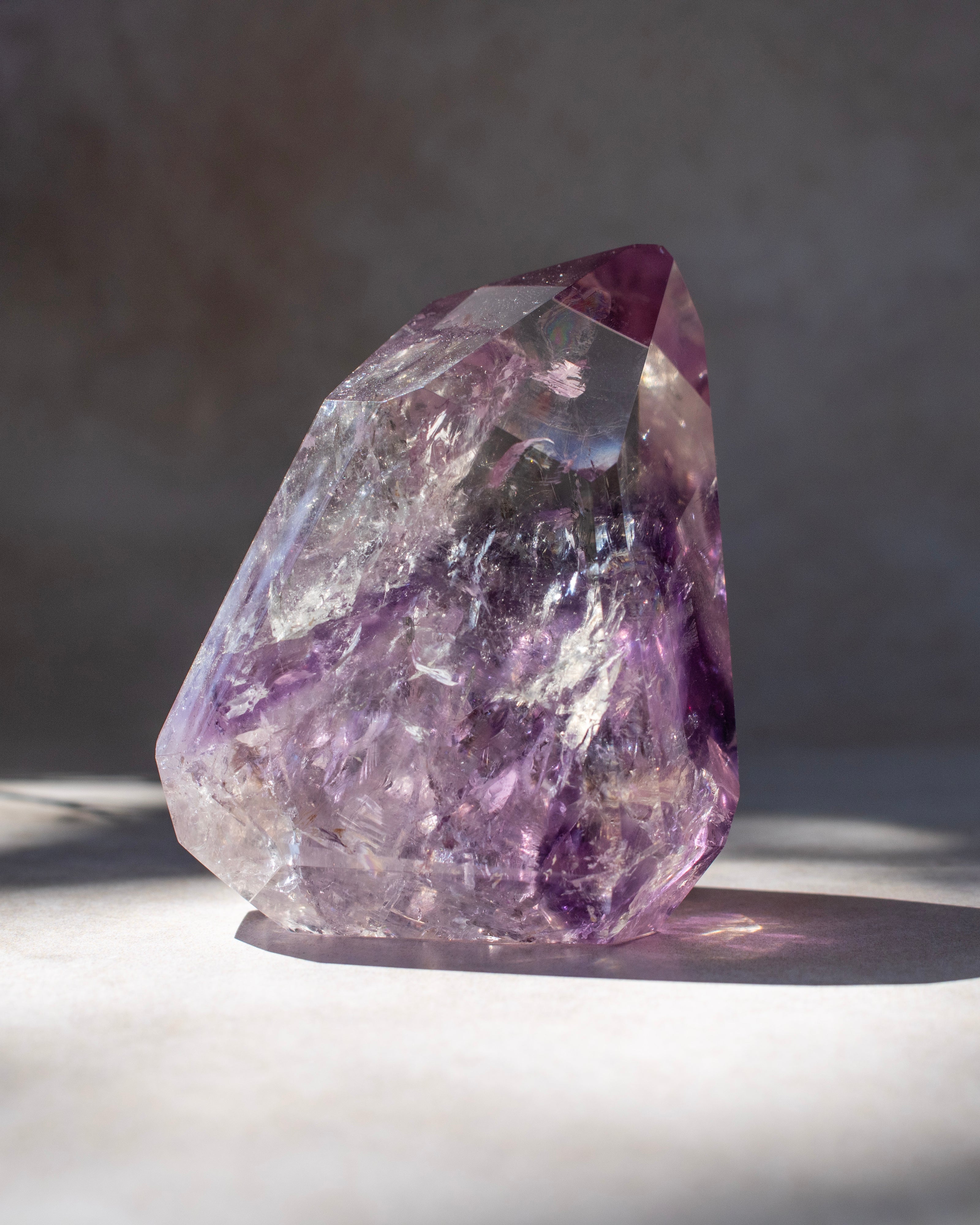 A raw purple amethyst crystal cluster with natural faceted points, standing on a light surface under direct sunlight with a soft shadow.