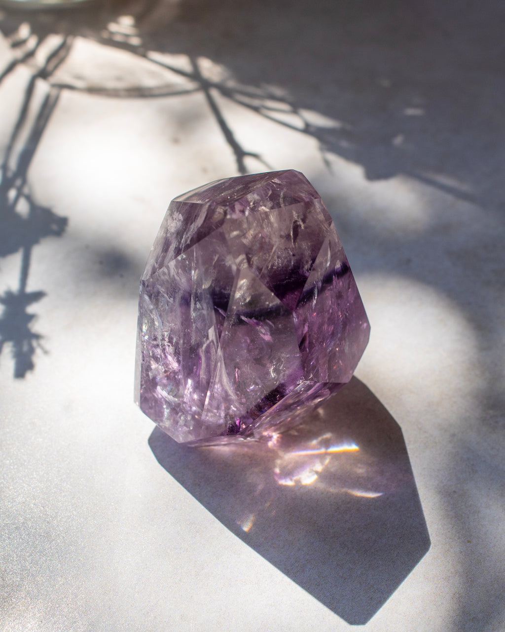 A faceted purple crystal gemstone, resembling amethyst, resting on a light gray surface with dappled sunlight and leaf shadows.