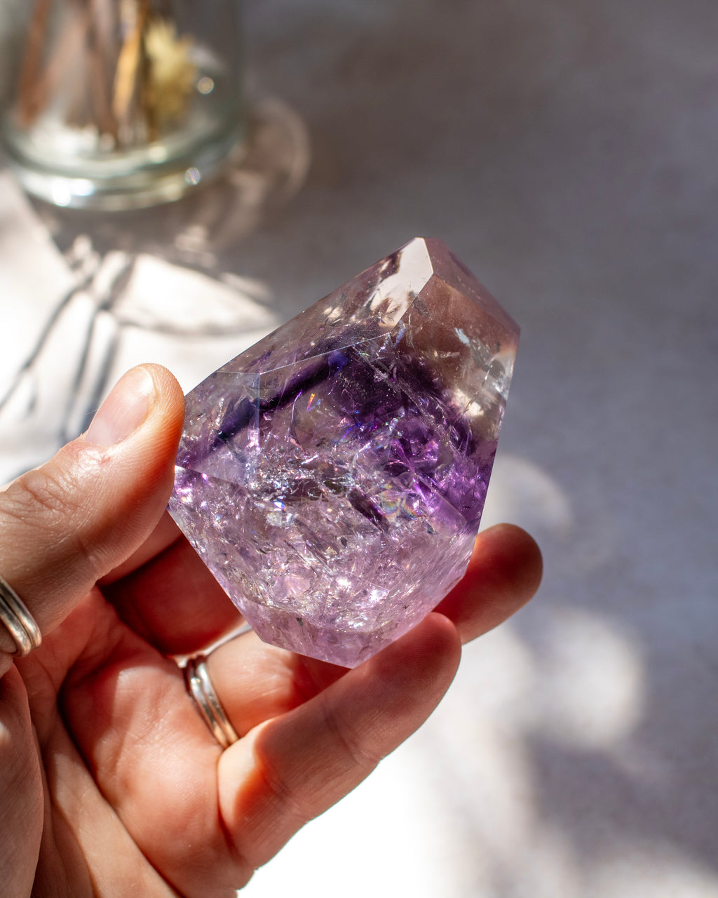 A hand holding a raw purple amethyst crystal point, with soft shadows and a blurred glass jar in the background.