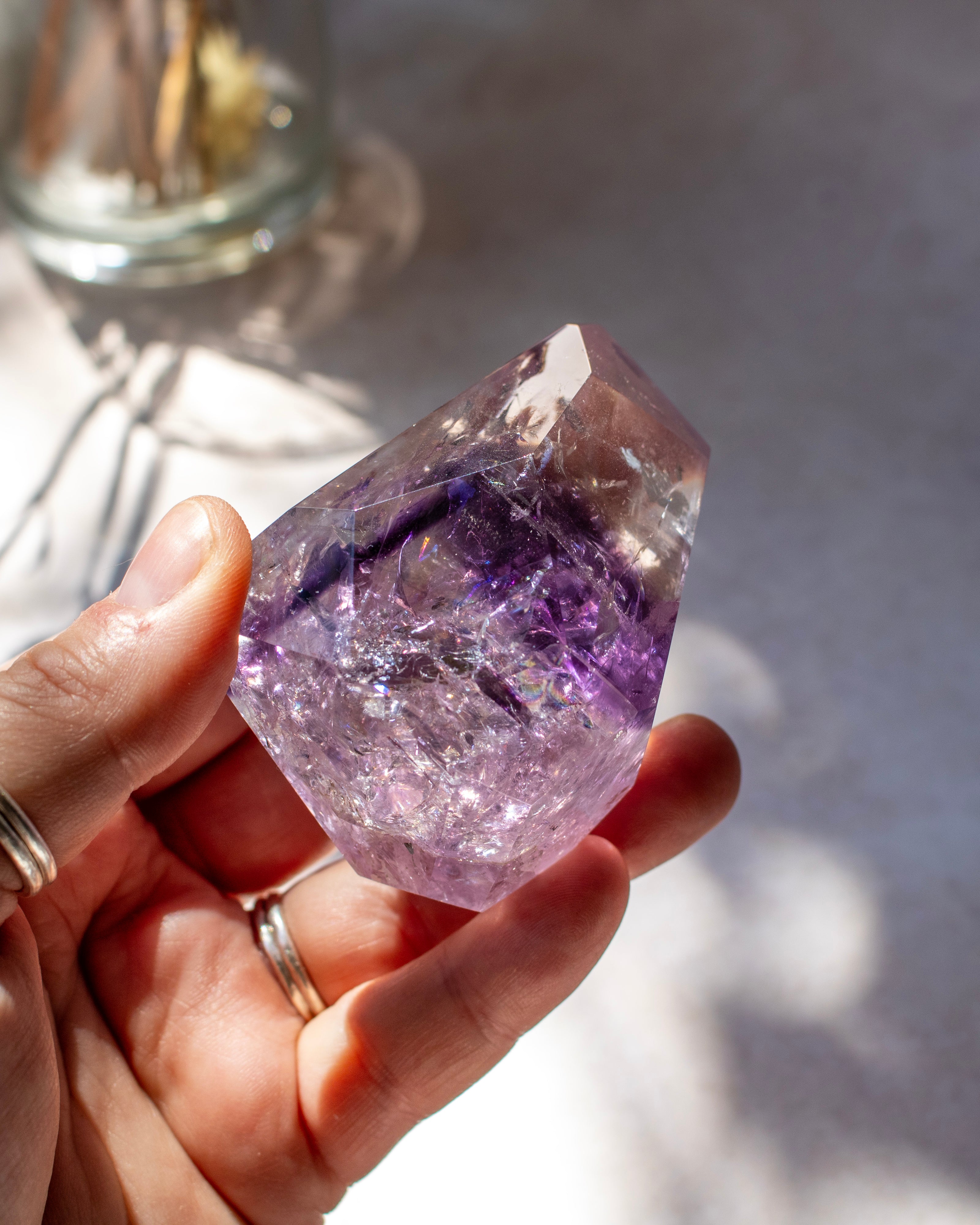 A hand holding a raw purple amethyst crystal point, with soft shadows and a blurred glass jar in the background.