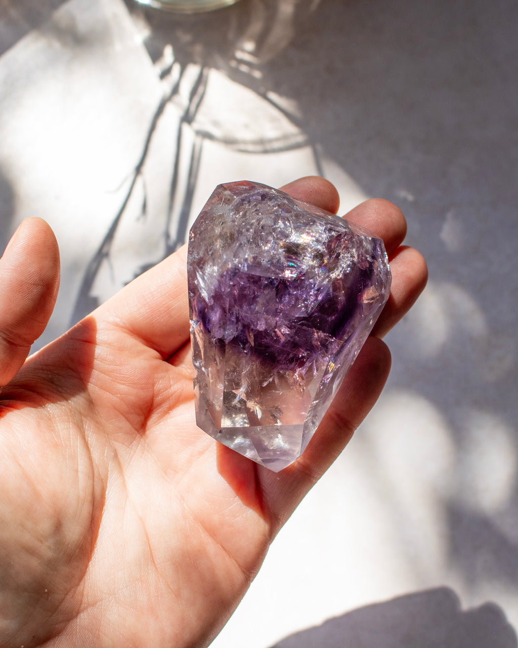 A hand holding a rough, unpolished amethyst crystal cluster with visible purple and clear layers, set against a bright white background with soft leaf shadows.