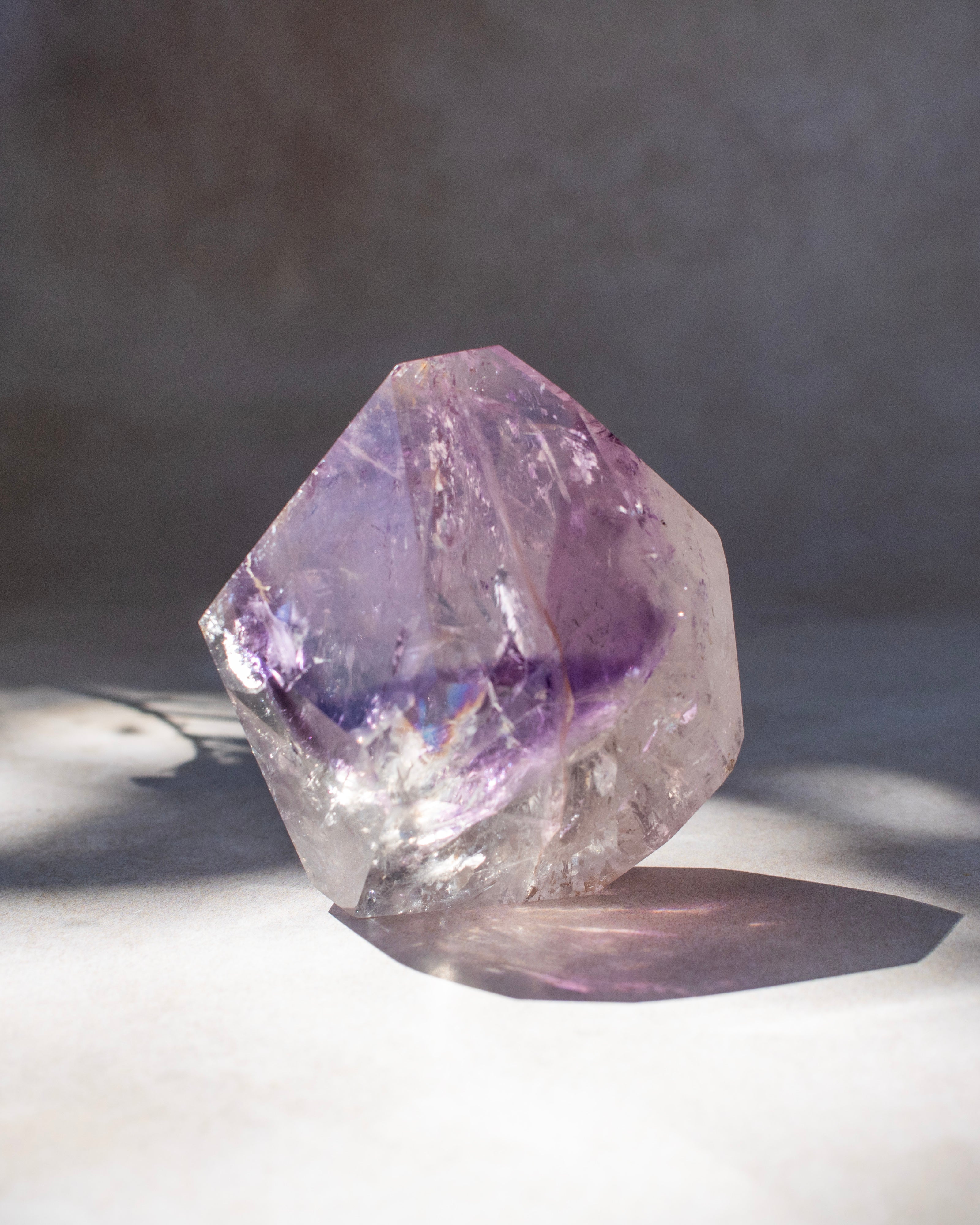 A raw amethyst crystal cluster with natural geometric facets, showing purple and clear quartz textures, resting on a light surface with a soft shadow.