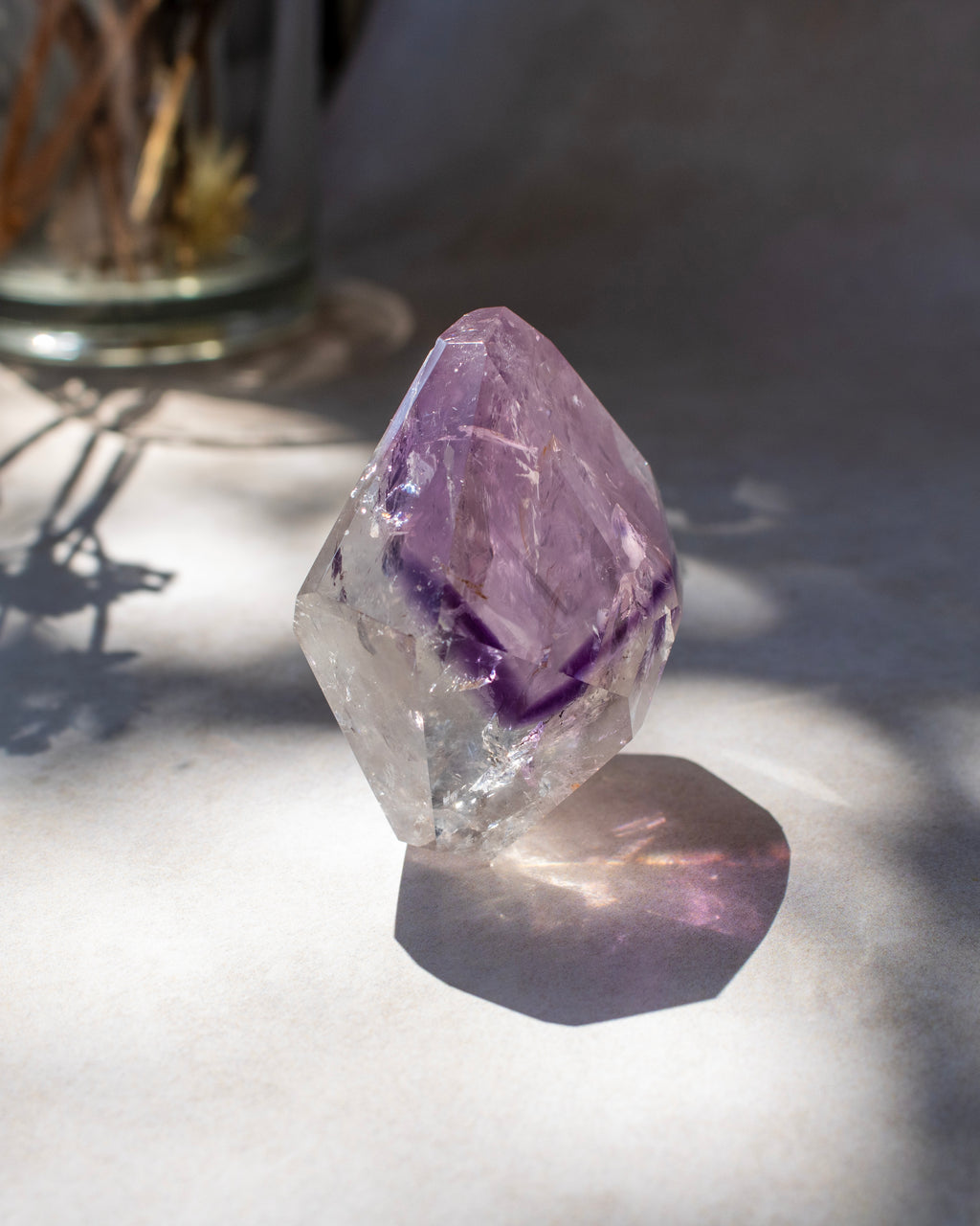 A raw amethyst crystal point with a clear faceted structure and a prominent purple inner core, resting on a light surface with dappled sunlight and shadows.