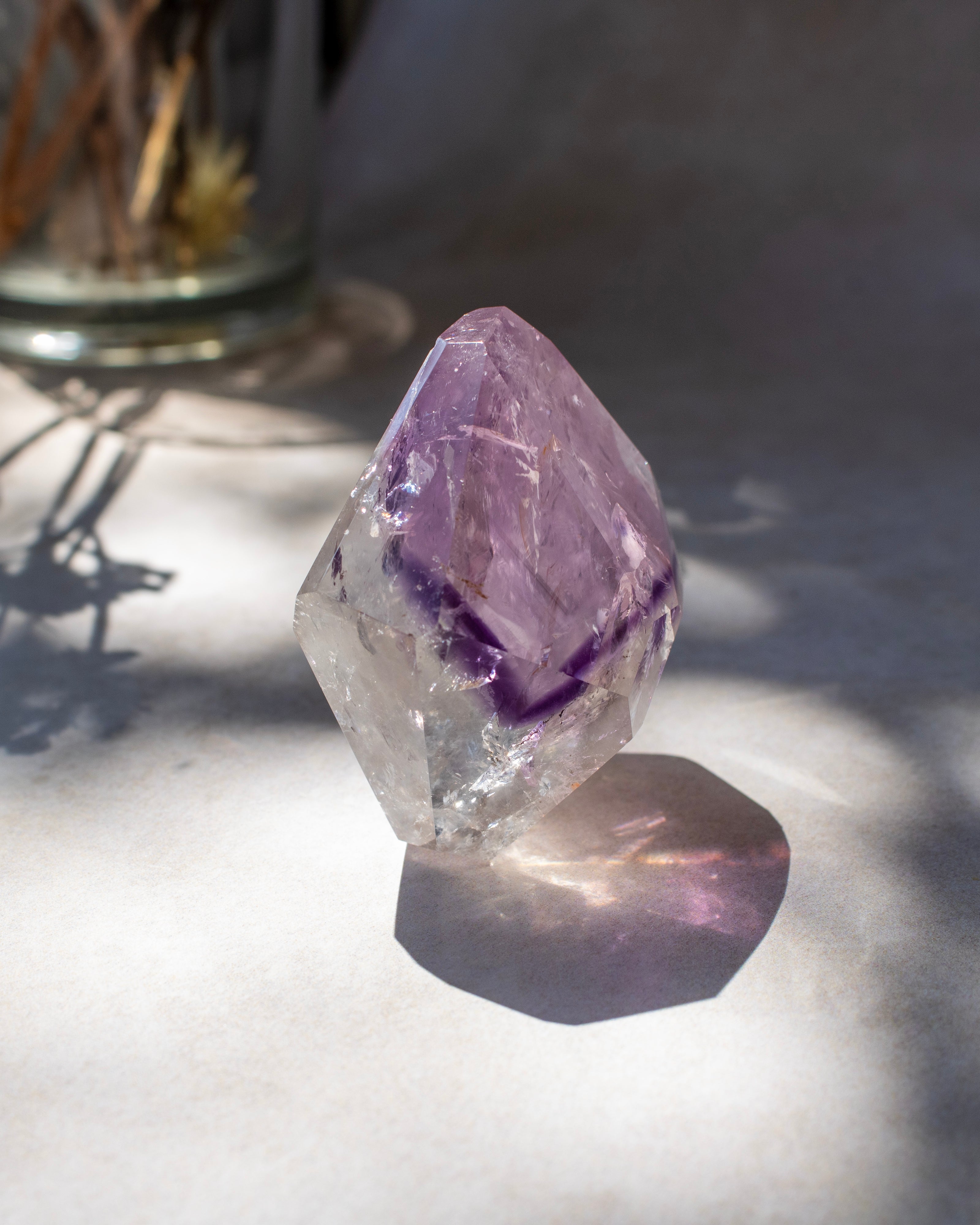 A raw amethyst crystal point with a clear faceted structure and a prominent purple inner core, resting on a light surface with dappled sunlight and shadows.