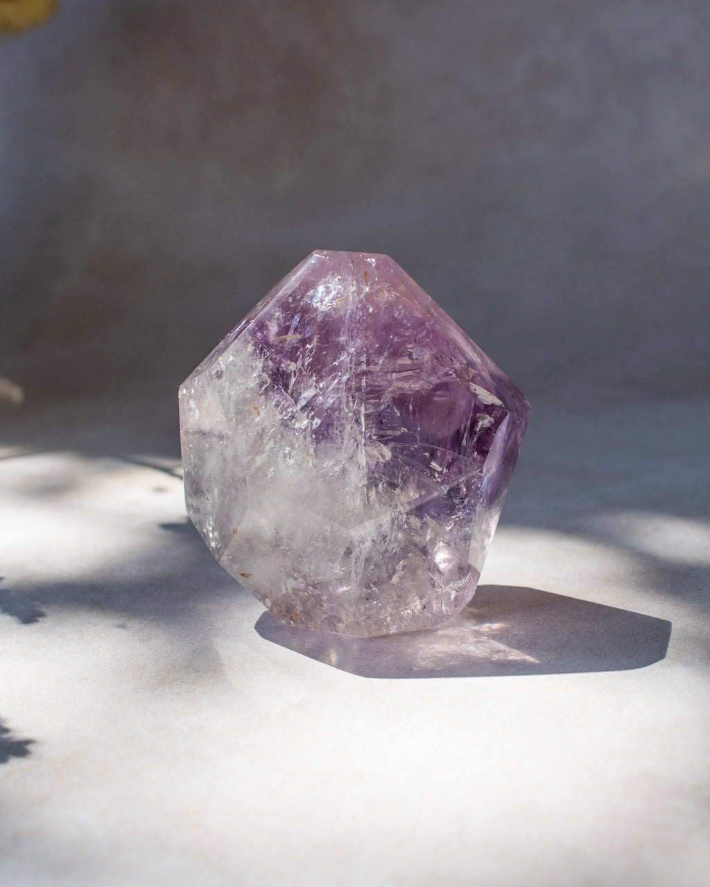 A raw amethyst crystal cluster with natural facets, showing purple and clear quartz textures, resting on a light surface with soft shadows.