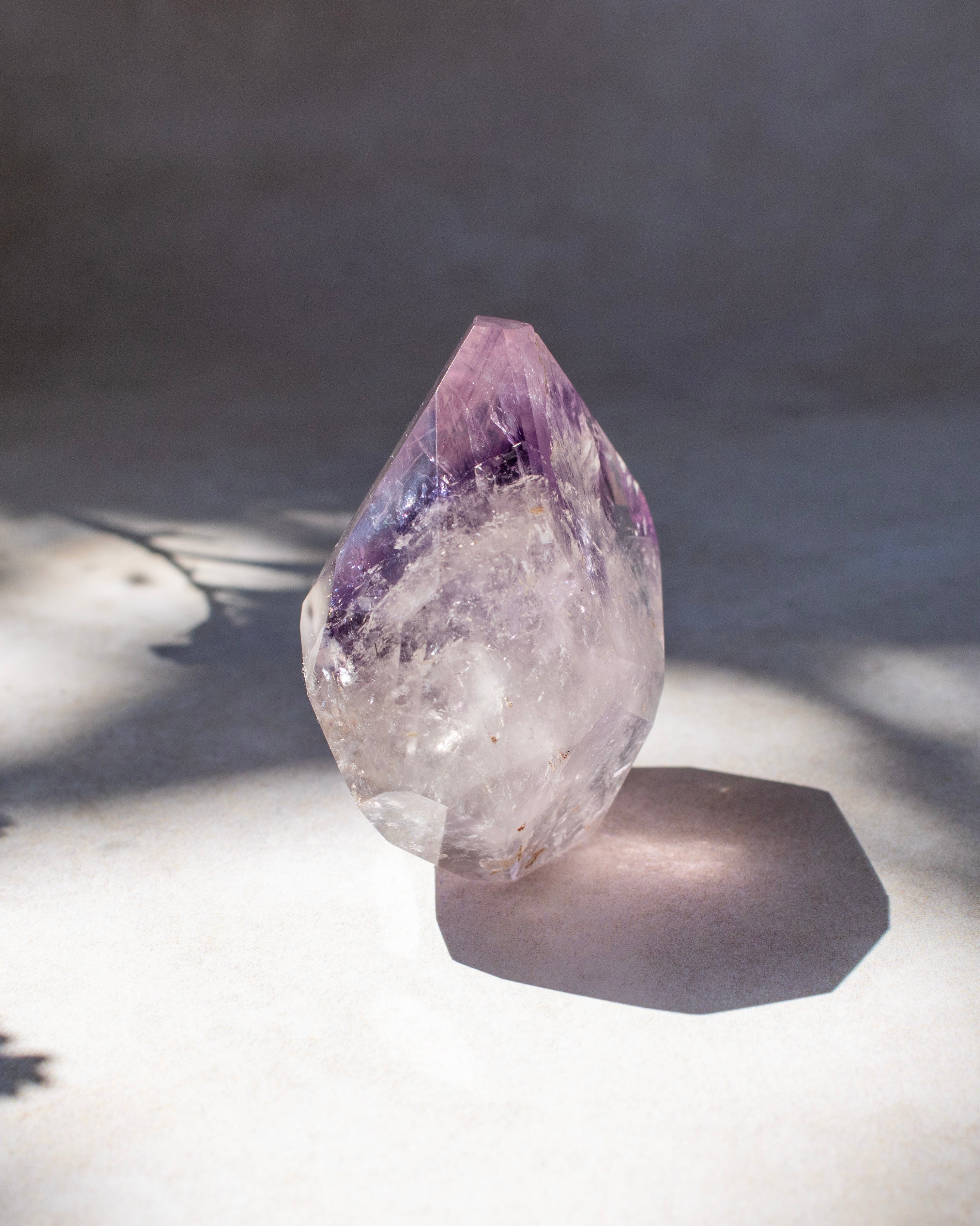 A raw amethyst crystal point with a purple top section and a clear, translucent lower body, standing on a light surface with a hard shadow under direct sunlight.