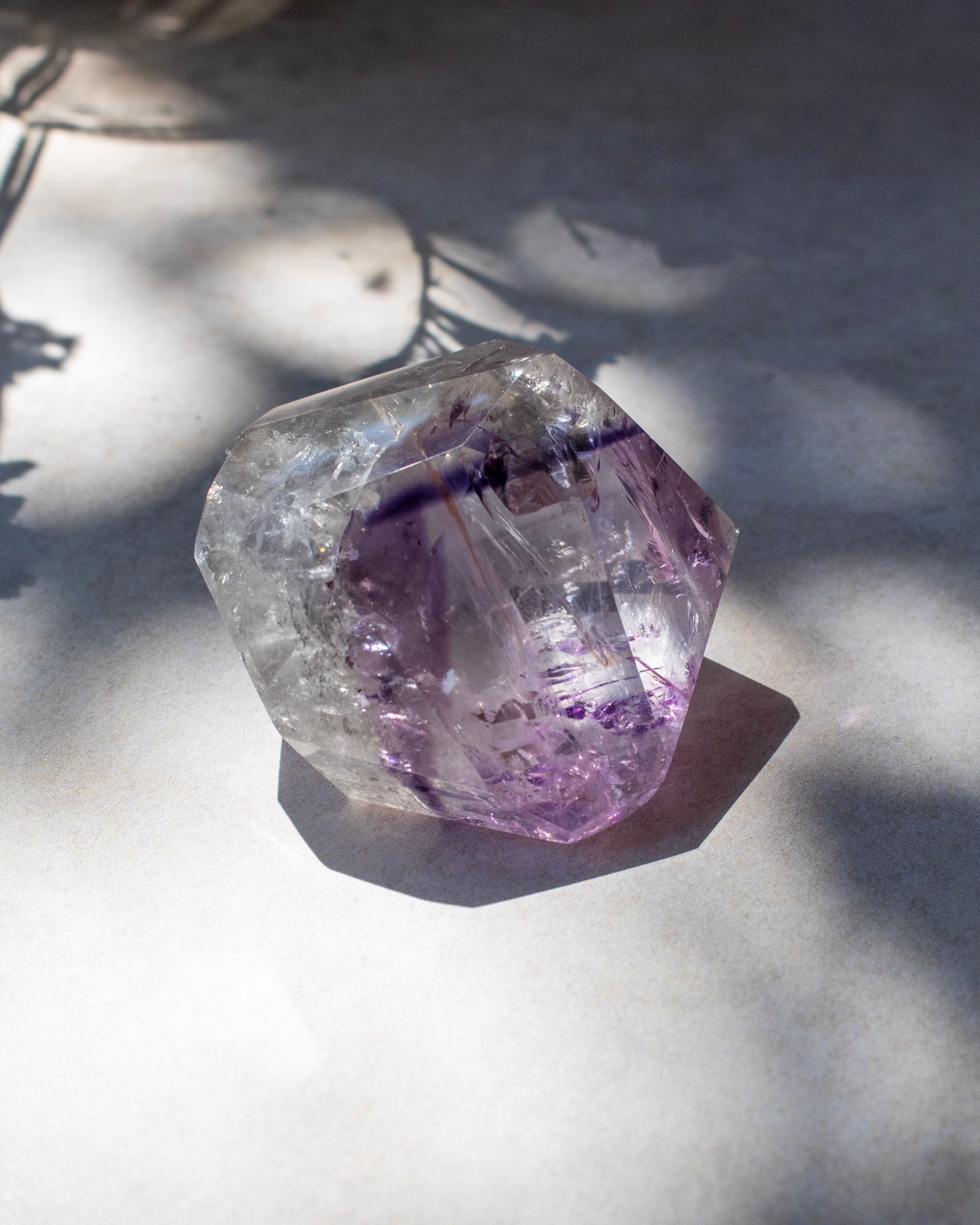A raw amethyst crystal cluster with purple and lavender tones, showing natural inclusions and faceted geometric edges, photographed on a white surface with dappled sunlight and shadows.