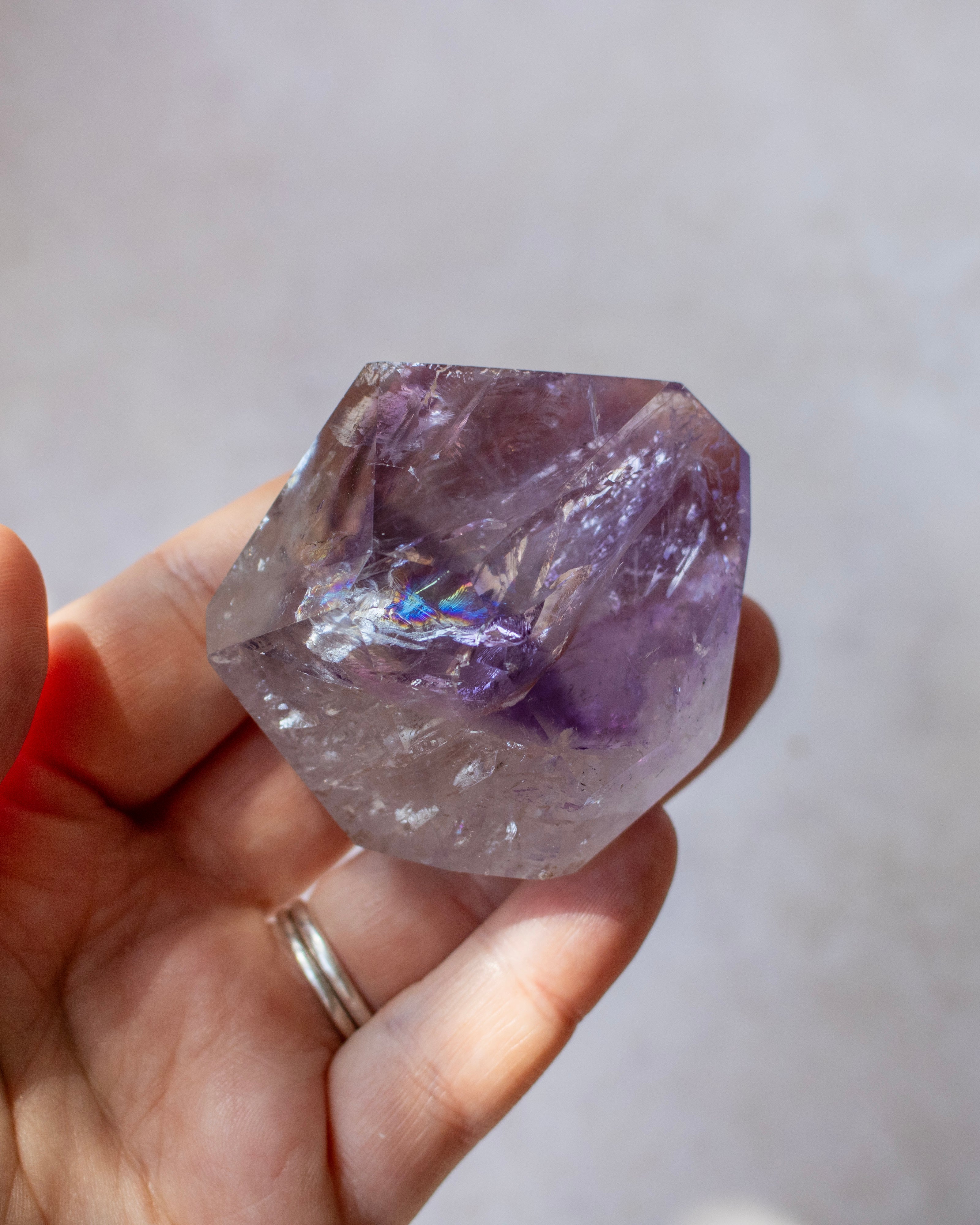A hand holding a faceted purple crystal gemstone with natural inclusions and a subtle iridescent sheen, set against a soft, out-of-focus light gray background.