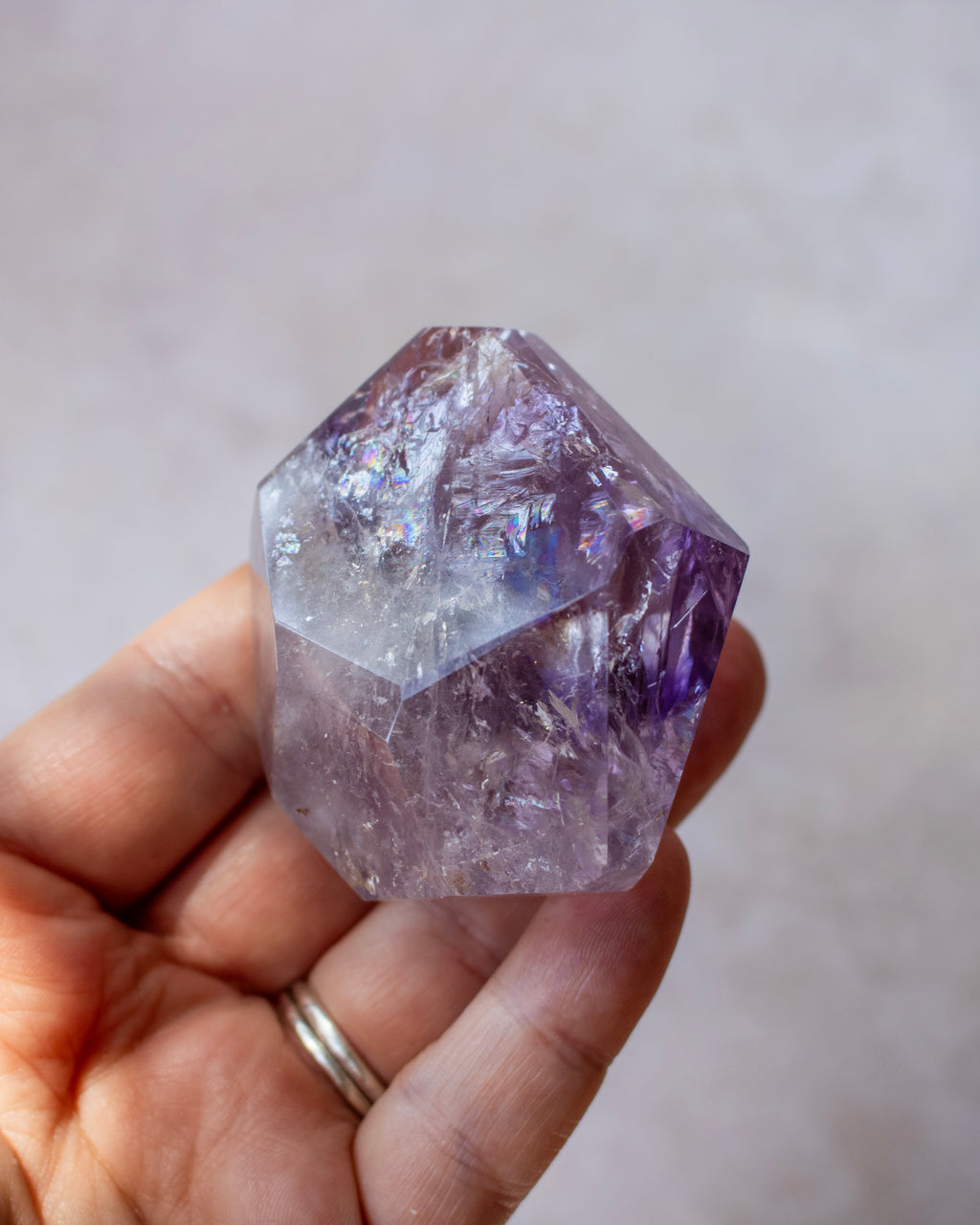 A hand holding a faceted purple amethyst crystal point against a soft, out-of-focus light gray background.