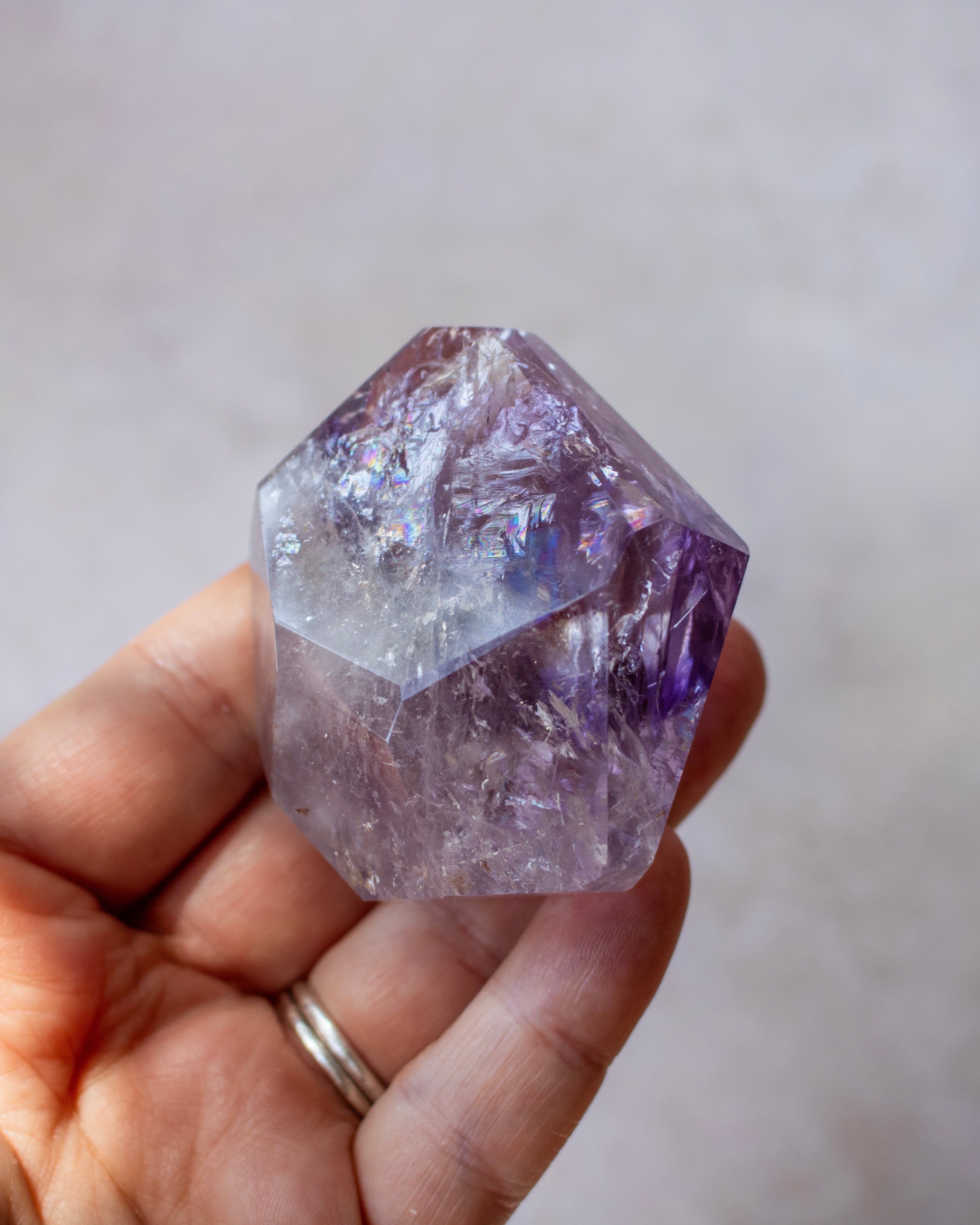 A hand holding a faceted purple amethyst crystal point against a soft, out-of-focus light gray background.