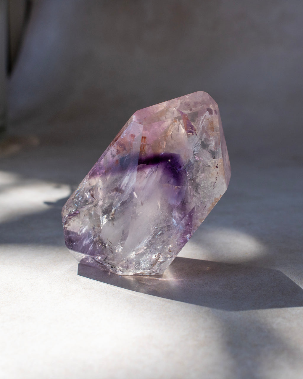 A raw, rough-cut amethyst crystal specimen with natural purple and clear sections, standing on a neutral surface in soft, natural light.
