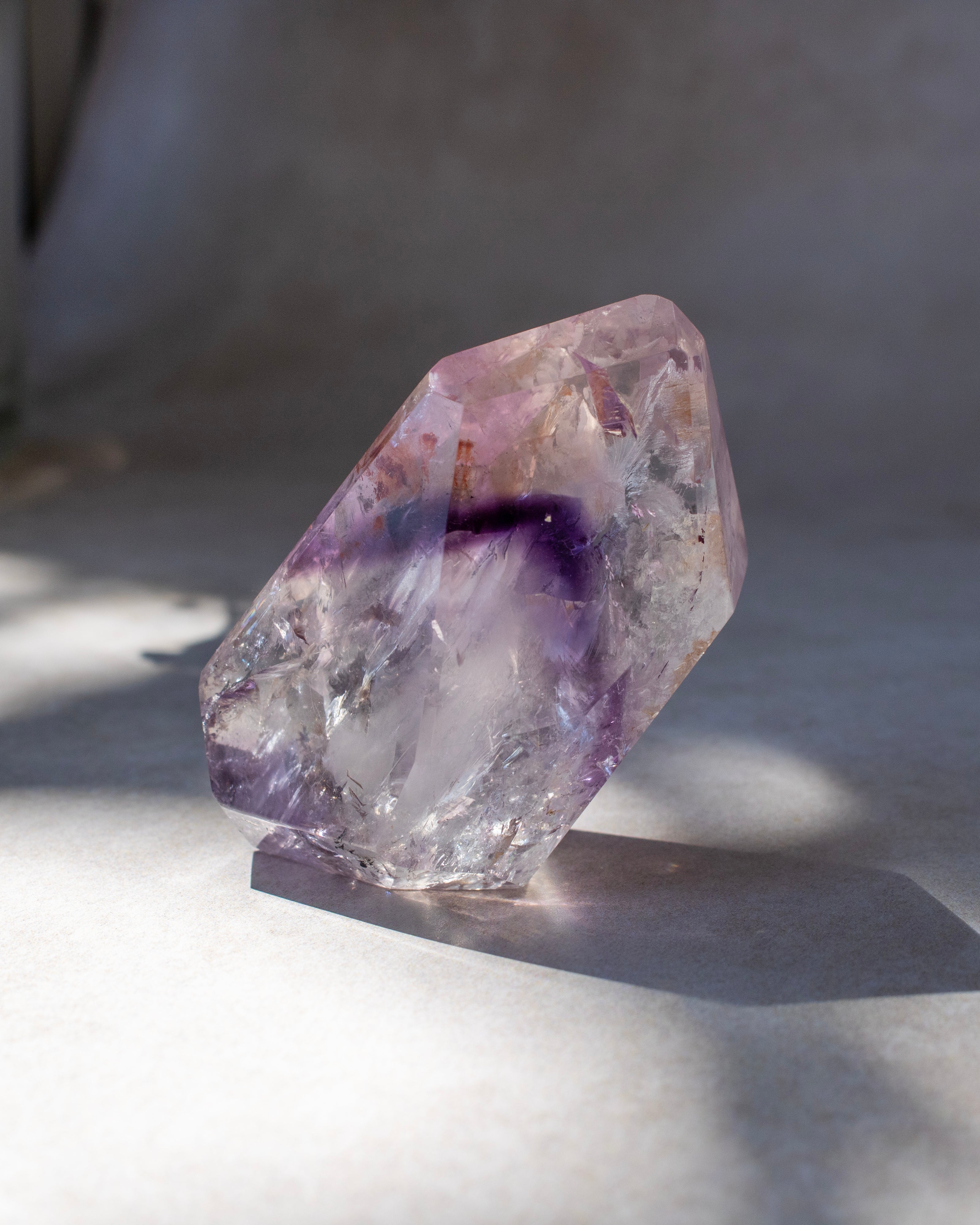 A raw, rough-cut amethyst crystal specimen with natural purple and clear sections, standing on a neutral surface in soft, natural light.