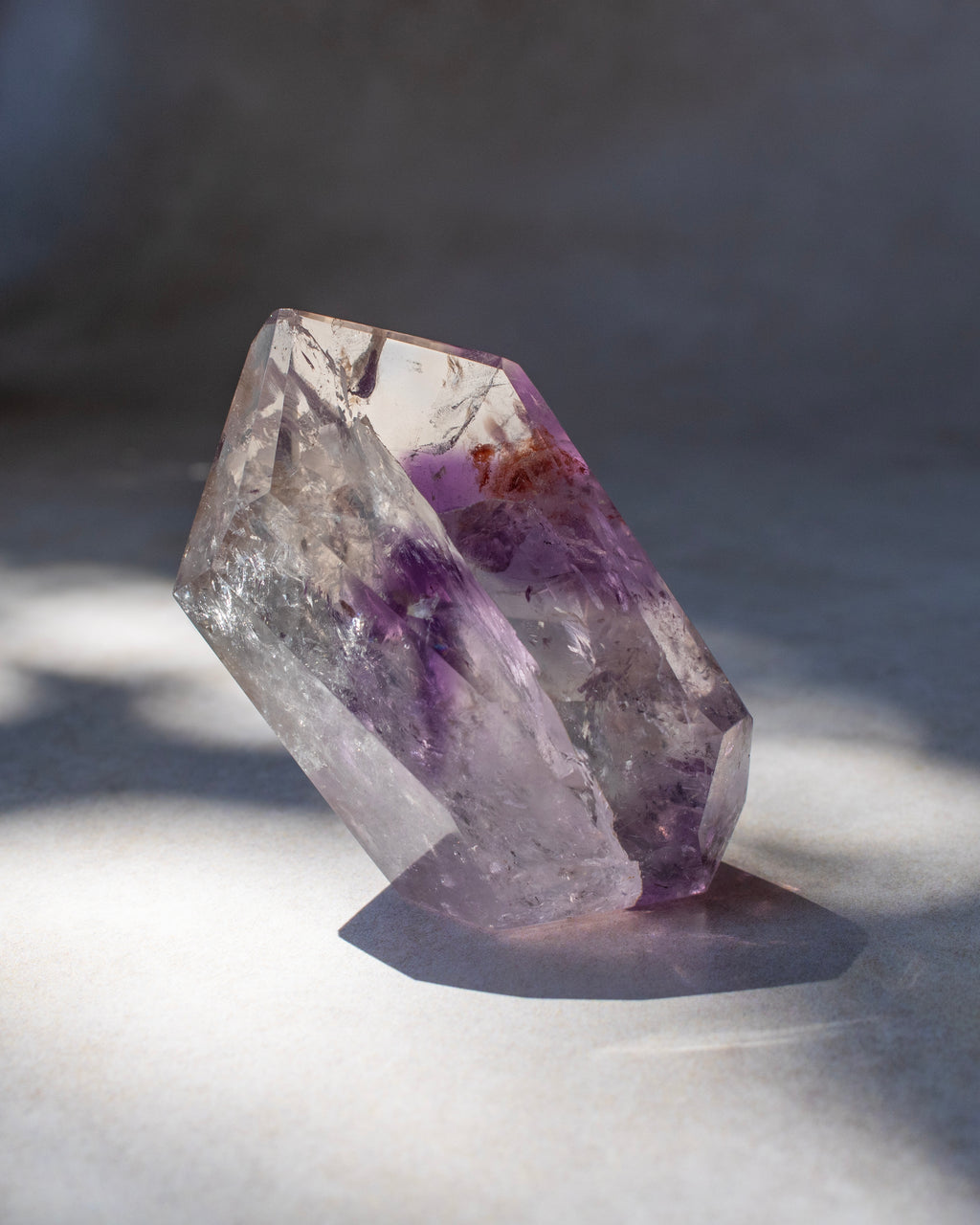 A natural purple amethyst crystal cluster with clear and lavender-purple sections, standing on a light gray surface with dramatic shadow lighting.