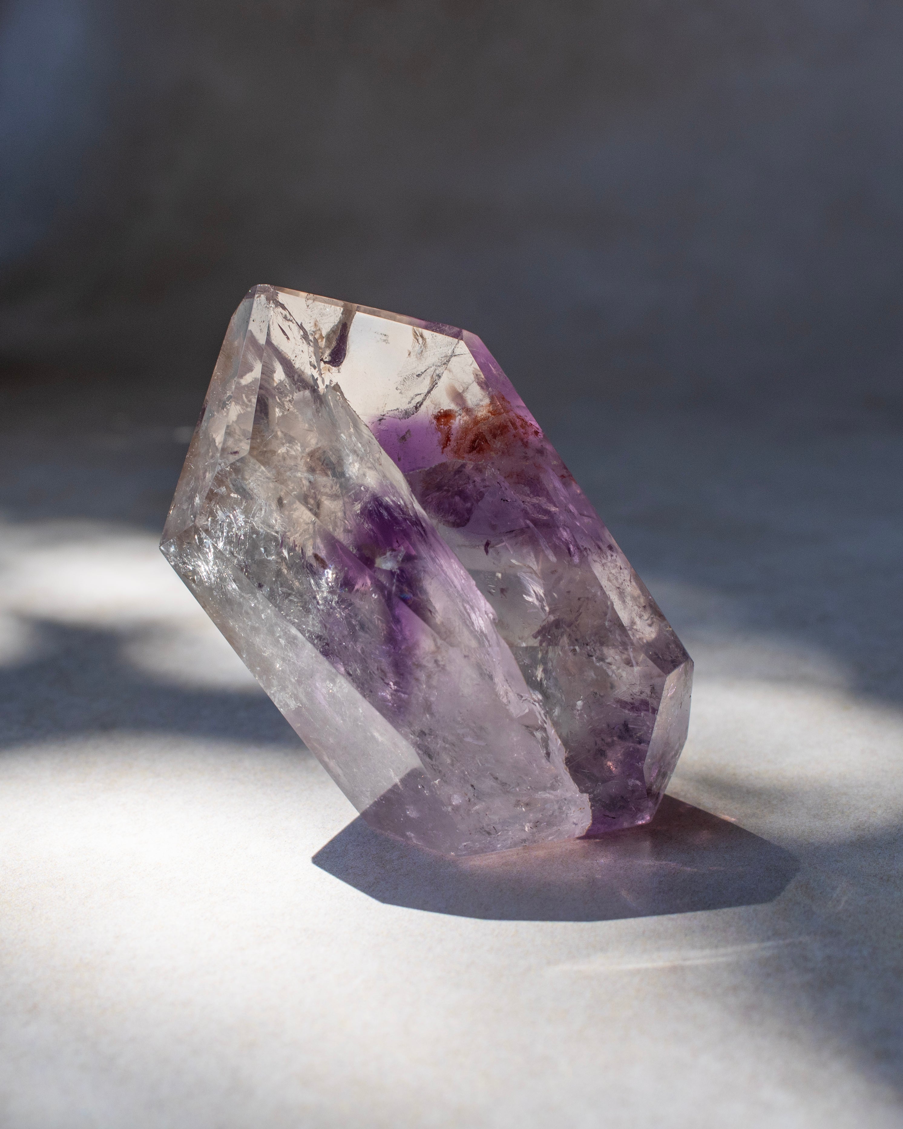 A natural purple amethyst crystal cluster with clear and lavender-purple sections, standing on a light gray surface with dramatic shadow lighting.
