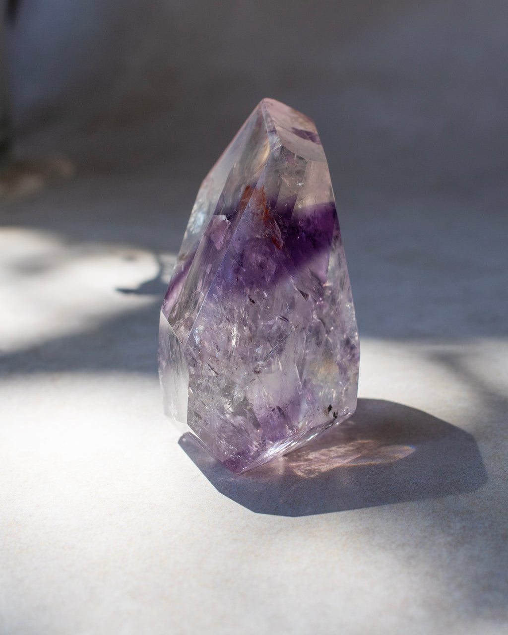 A natural clear quartz crystal point with prominent purple amethyst inclusions, standing on a light gray surface under direct sunlight.