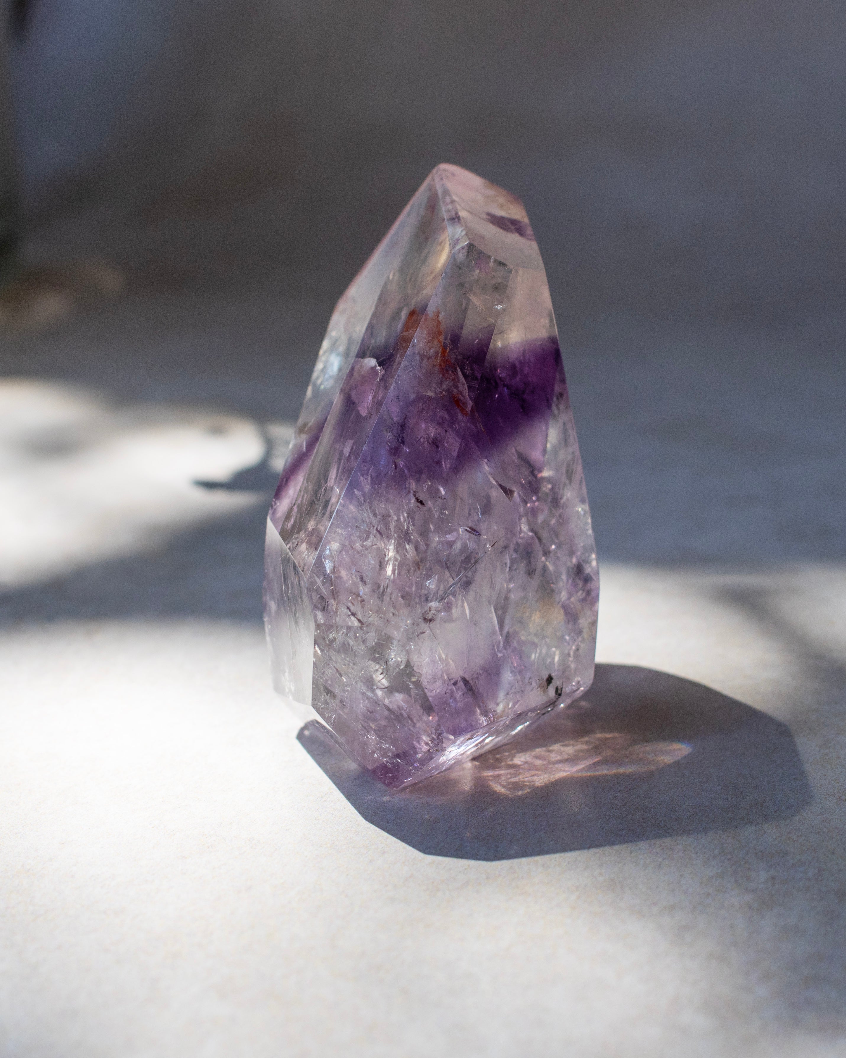 A natural clear quartz crystal point with prominent purple amethyst inclusions, standing on a light gray surface under direct sunlight.