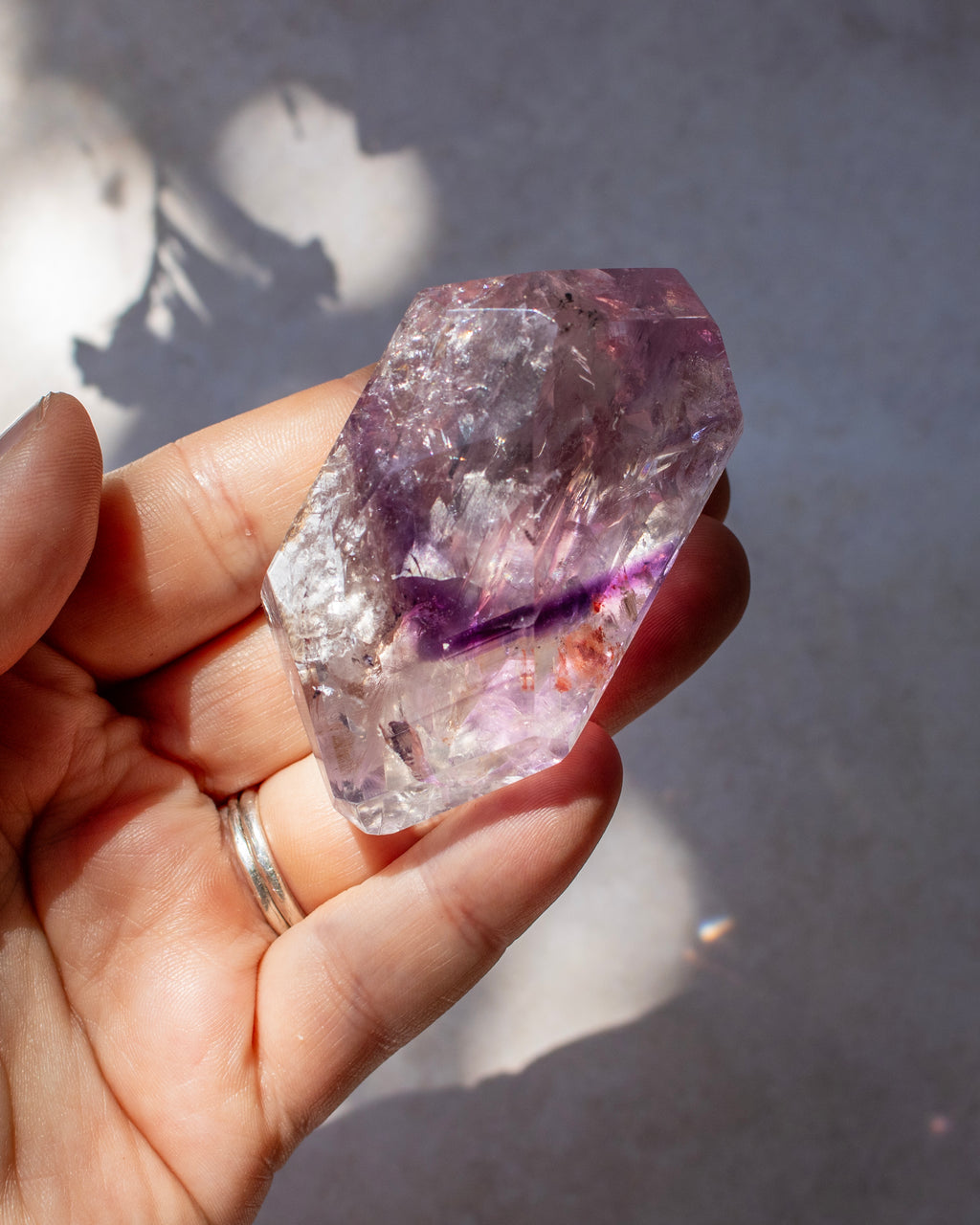 A hand holding a faceted purple amethyst crystal point under natural sunlight against a gray background with soft shadows.