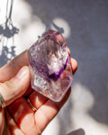 A hand holding a raw amethyst crystal point with natural purple and clear quartz formations, photographed outdoors in bright sunlight with soft shadows.