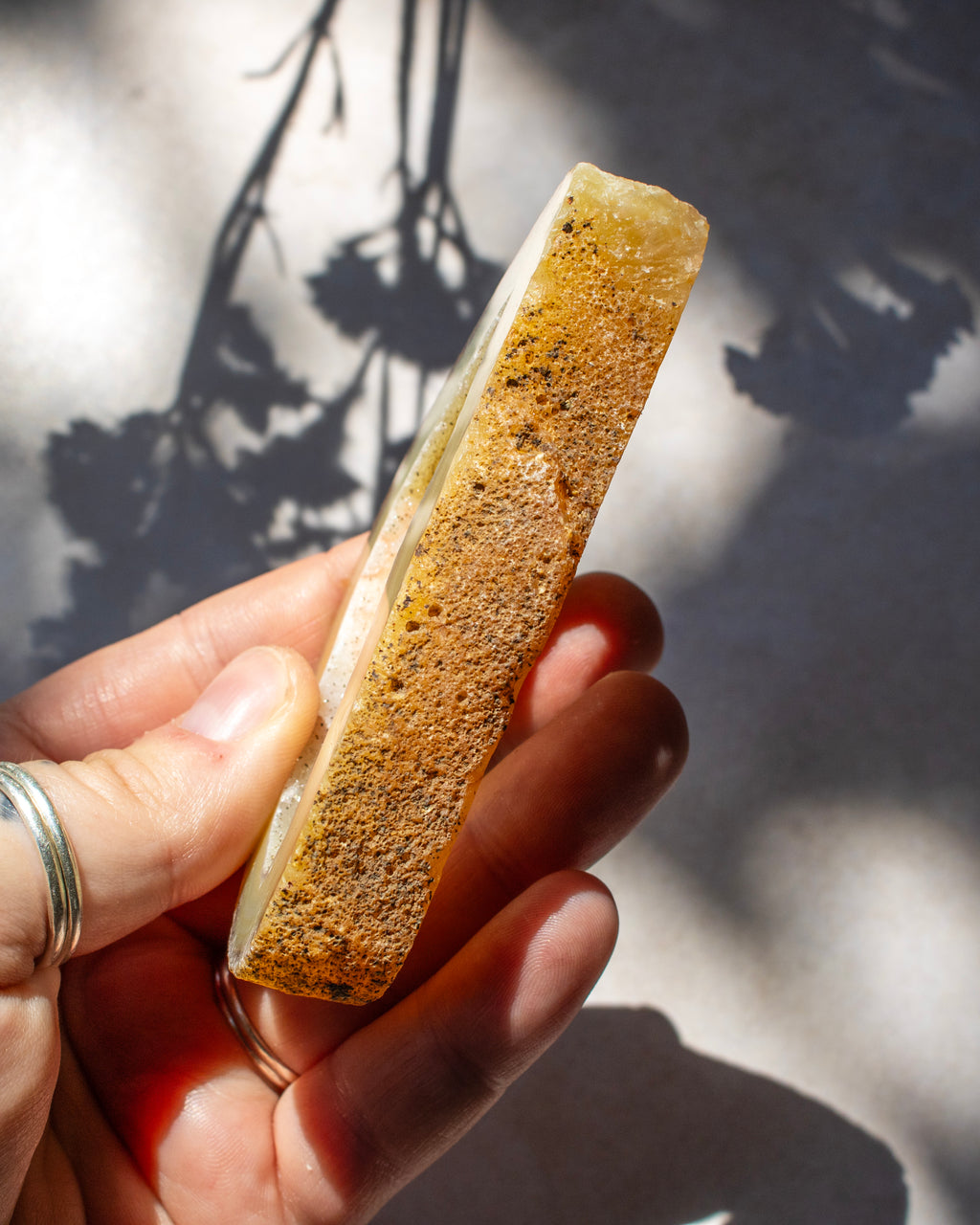 A hand holding a rectangular natural crystal or mineral specimen with a rough, porous golden-brown texture and a pale translucent edge, photographed outdoors with strong sunlight and leaf shadows on a light surface.