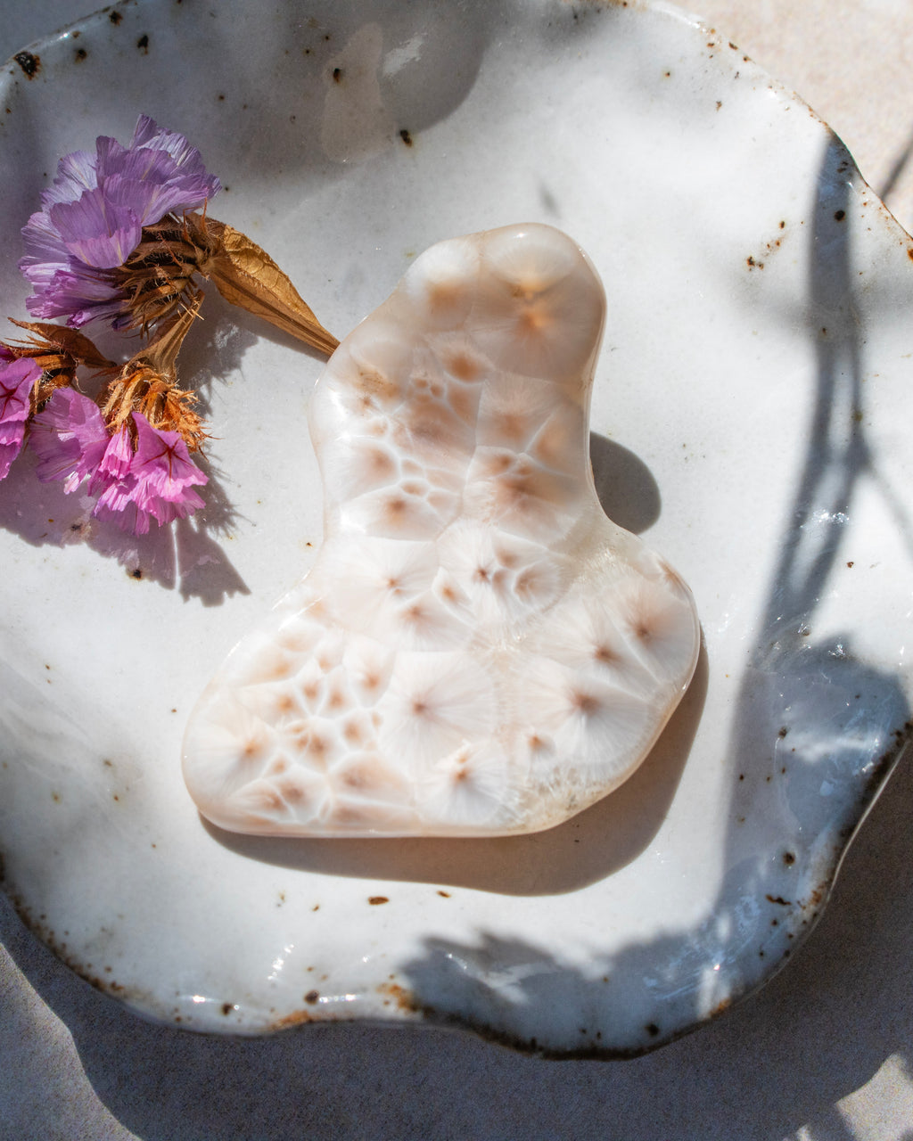 A carved white stone or bone figurine with organic, porous patterns, displayed on a rustic white speckled ceramic plate with dried purple flowers under natural sunlight.