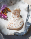 A carved white stone or bone figurine with organic, porous patterns, displayed on a rustic white speckled ceramic plate with dried purple flowers under natural sunlight.