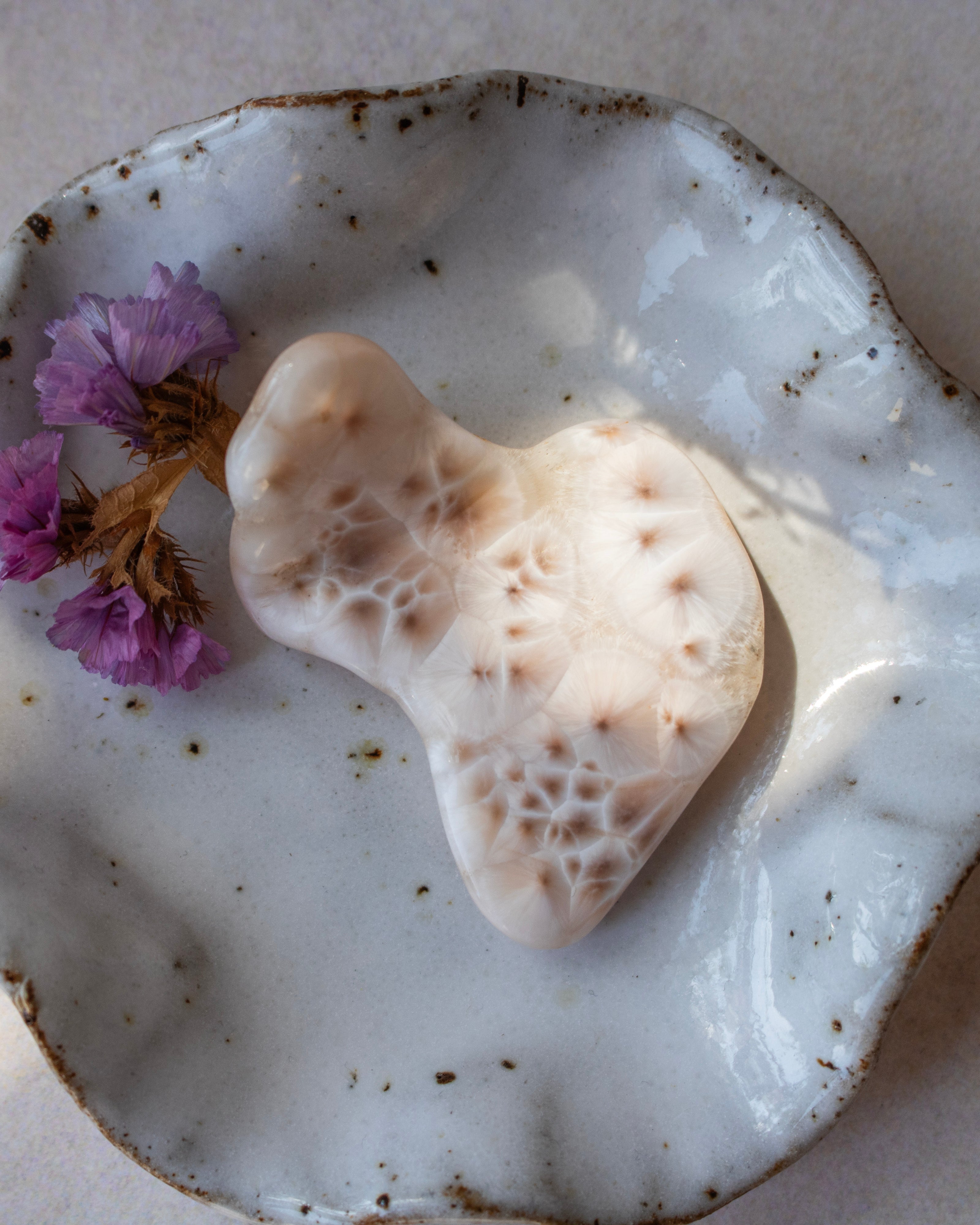 A heart-shaped polished stone with intricate web-like patterns, displayed on a rustic white speckled ceramic plate with dried purple flowers.