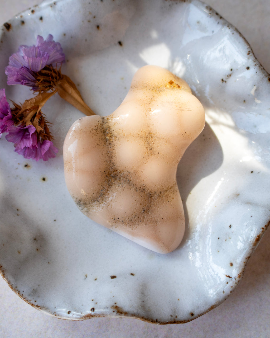 A polished beige stone with a bubbly, pearly texture sits on a white speckled ceramic dish with dried purple flowers as props.