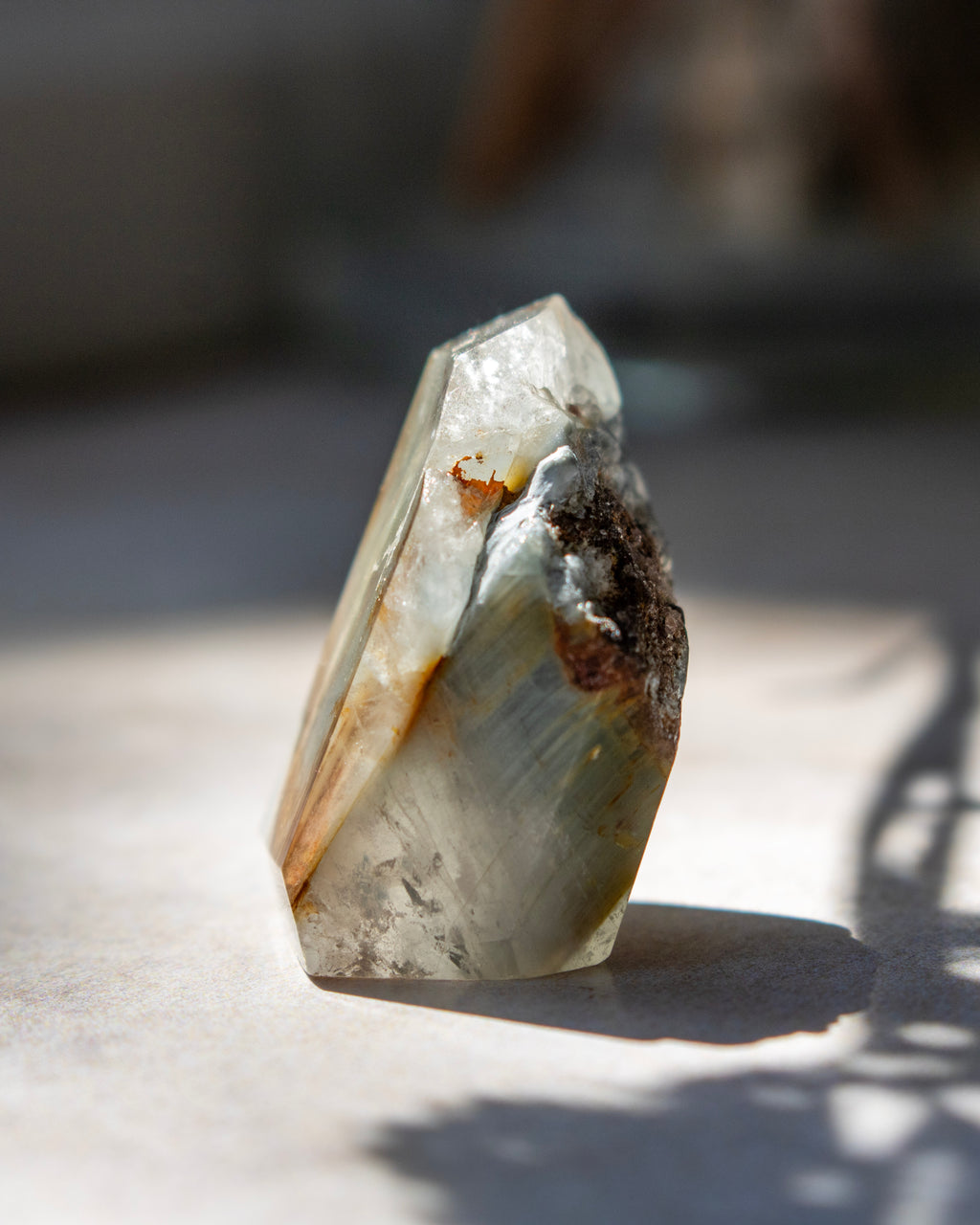 A raw crystal mineral specimen with a pointed tip, showing natural striations in pale gray, white, and brown, photographed on a light surface with dappled sunlight and shadows.