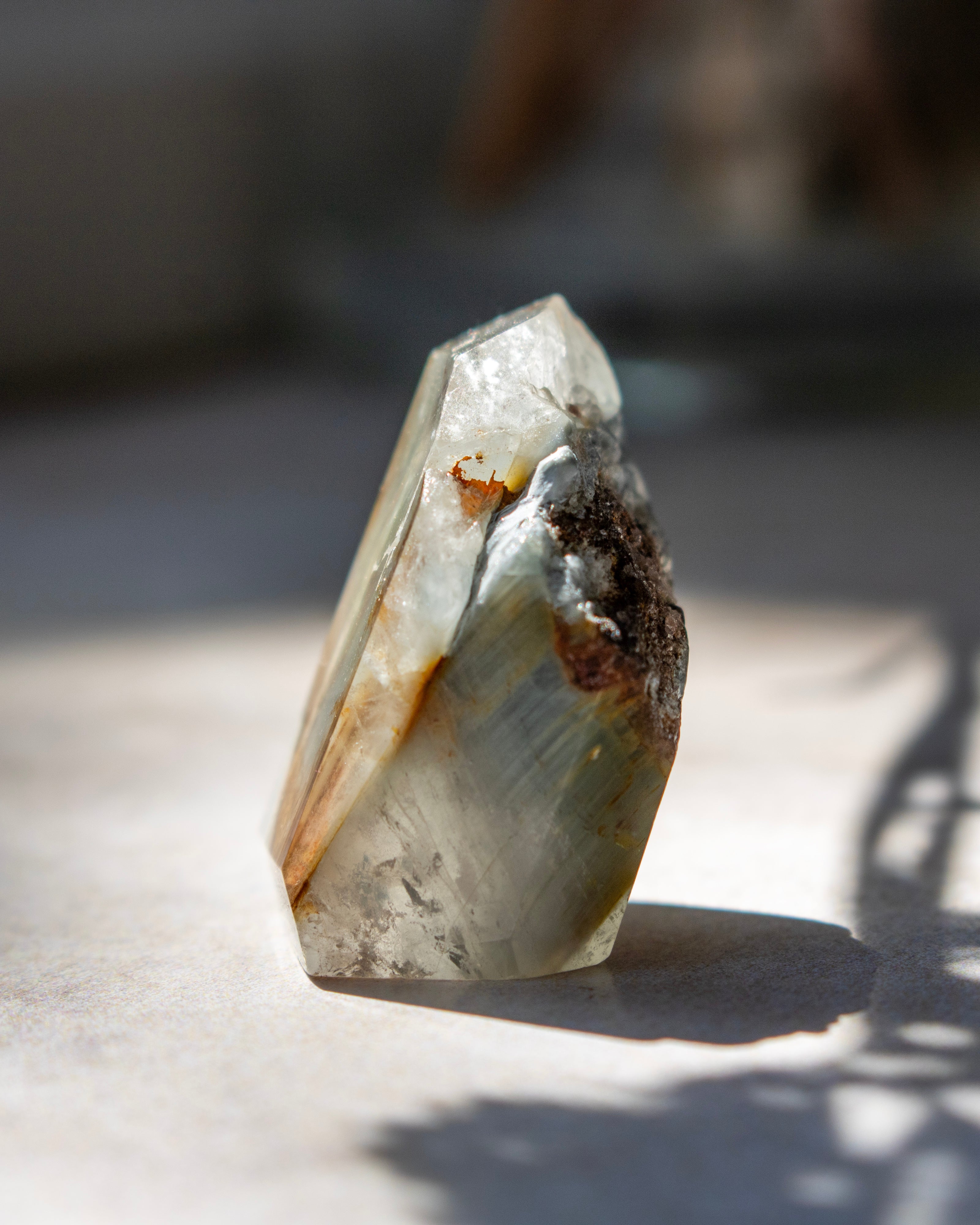 A raw crystal mineral specimen with a pointed tip, showing natural striations in pale gray, white, and brown, photographed on a light surface with dappled sunlight and shadows.