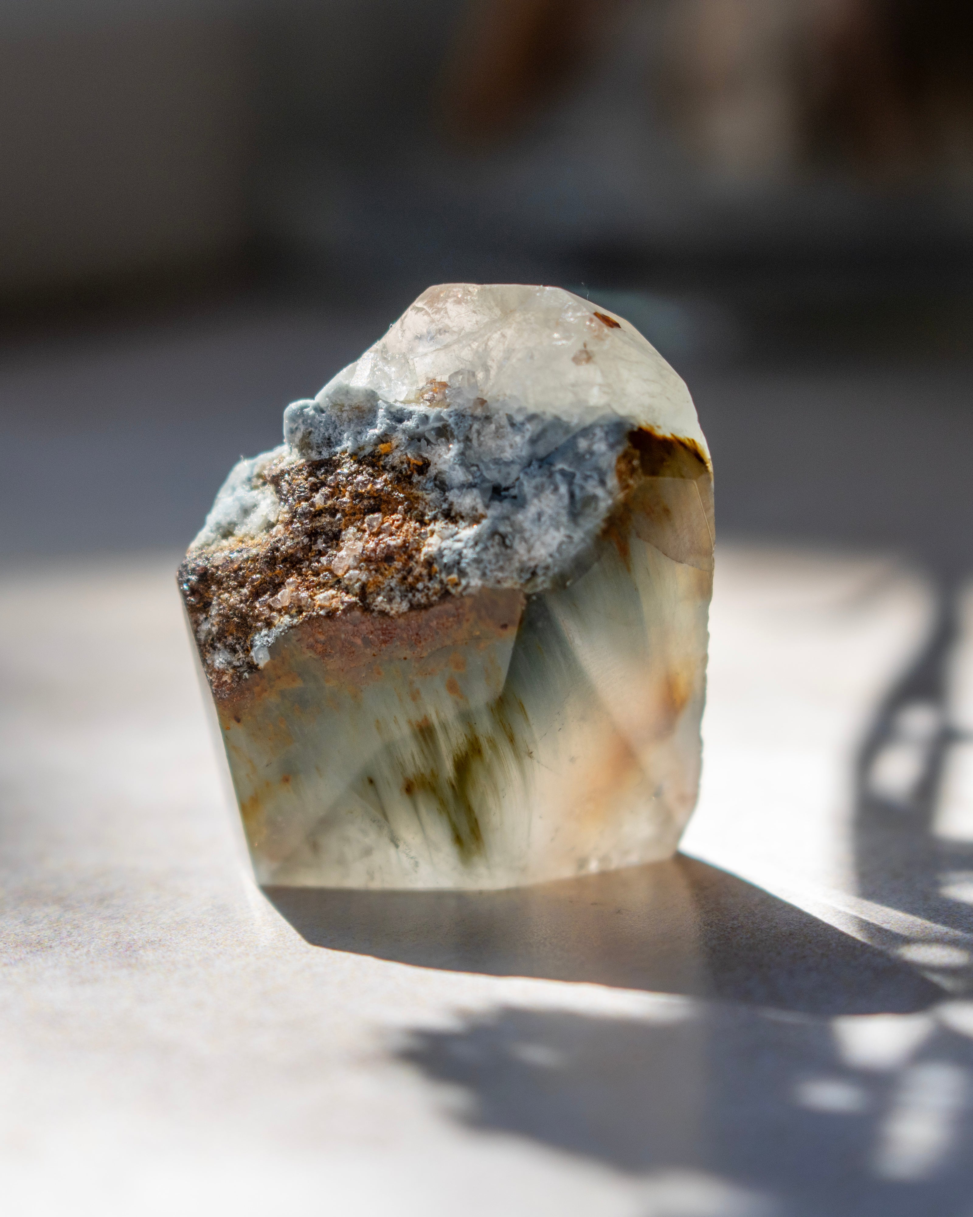 A raw, unpolished crystal mineral specimen with layered translucent bands of white, gray, and brown, standing on a light surface in direct sunlight.