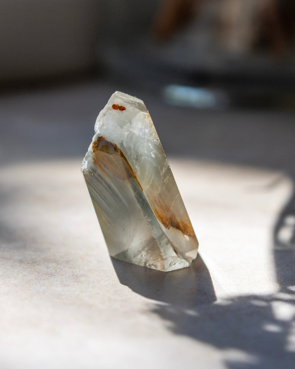 A natural clear quartz crystal point with internal brown and white inclusions, standing upright on a light surface with a soft shadow.