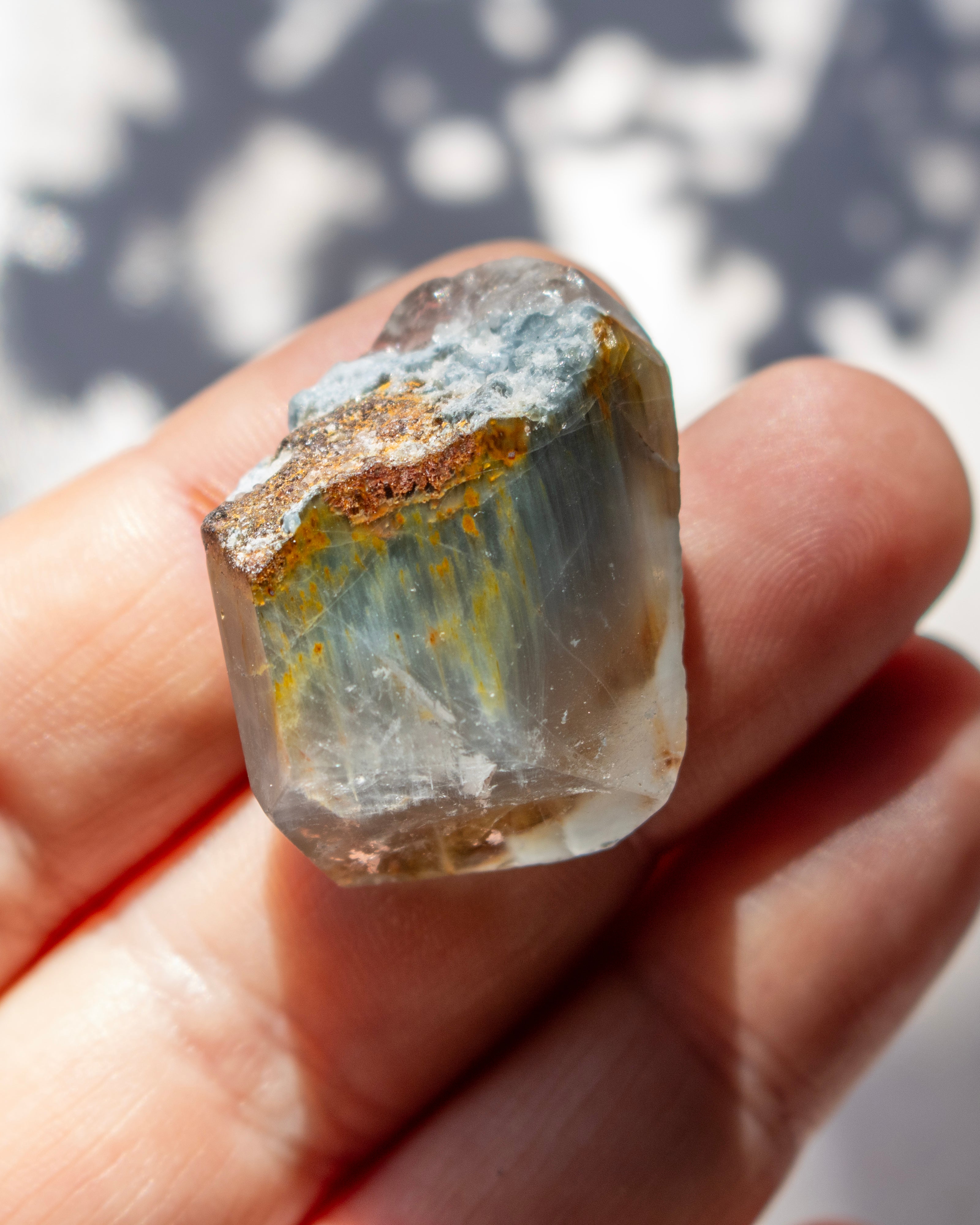 A hand holding a raw, rough gemstone with a clear body, yellow-green veining, and a metallic gold and brown crust.