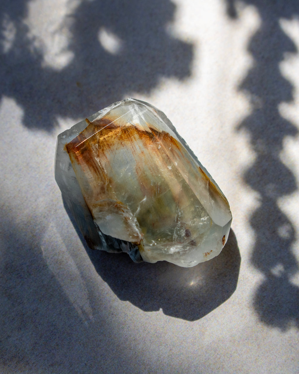 A raw, rough crystal cluster with natural brown and greenish-brown inclusions, resting on a light gray surface under dappled sunlight and shadows.
