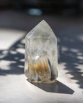A clear pointed crystal tower with internal amber and dark inclusions, photographed on a light surface in direct sunlight with strong shadows.