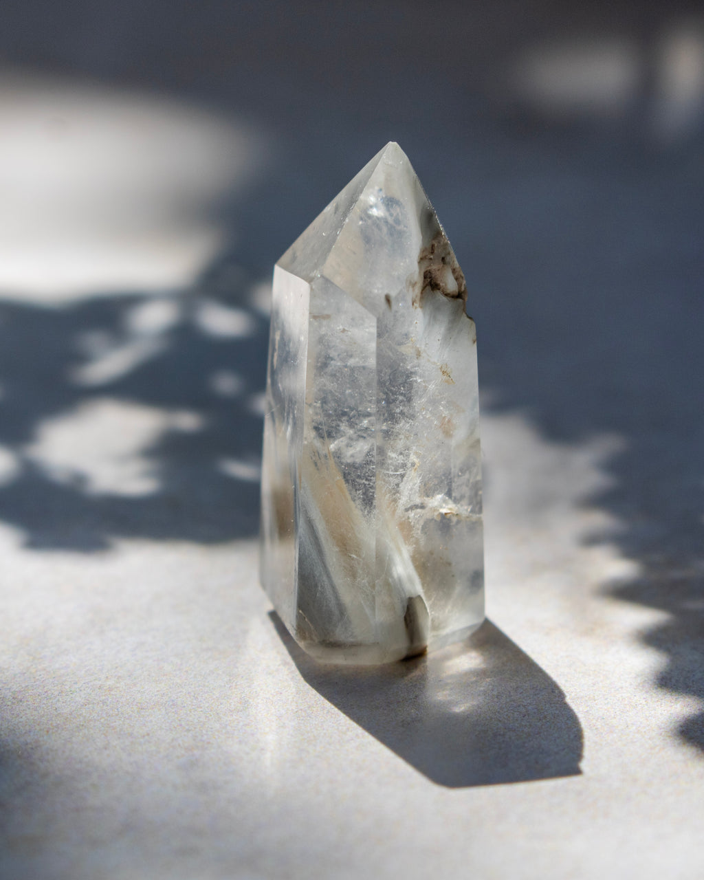 A natural clear quartz crystal point with inclusions, standing upright on a light surface under direct sunlight with leaf shadows.