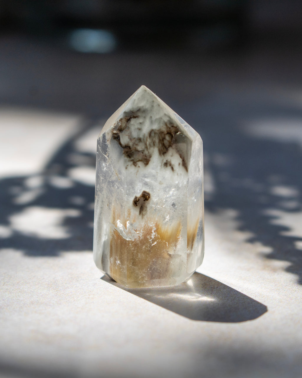A natural clear quartz crystal point with brown and tan inclusions, standing upright on a light surface in direct sunlight with a sharp shadow.