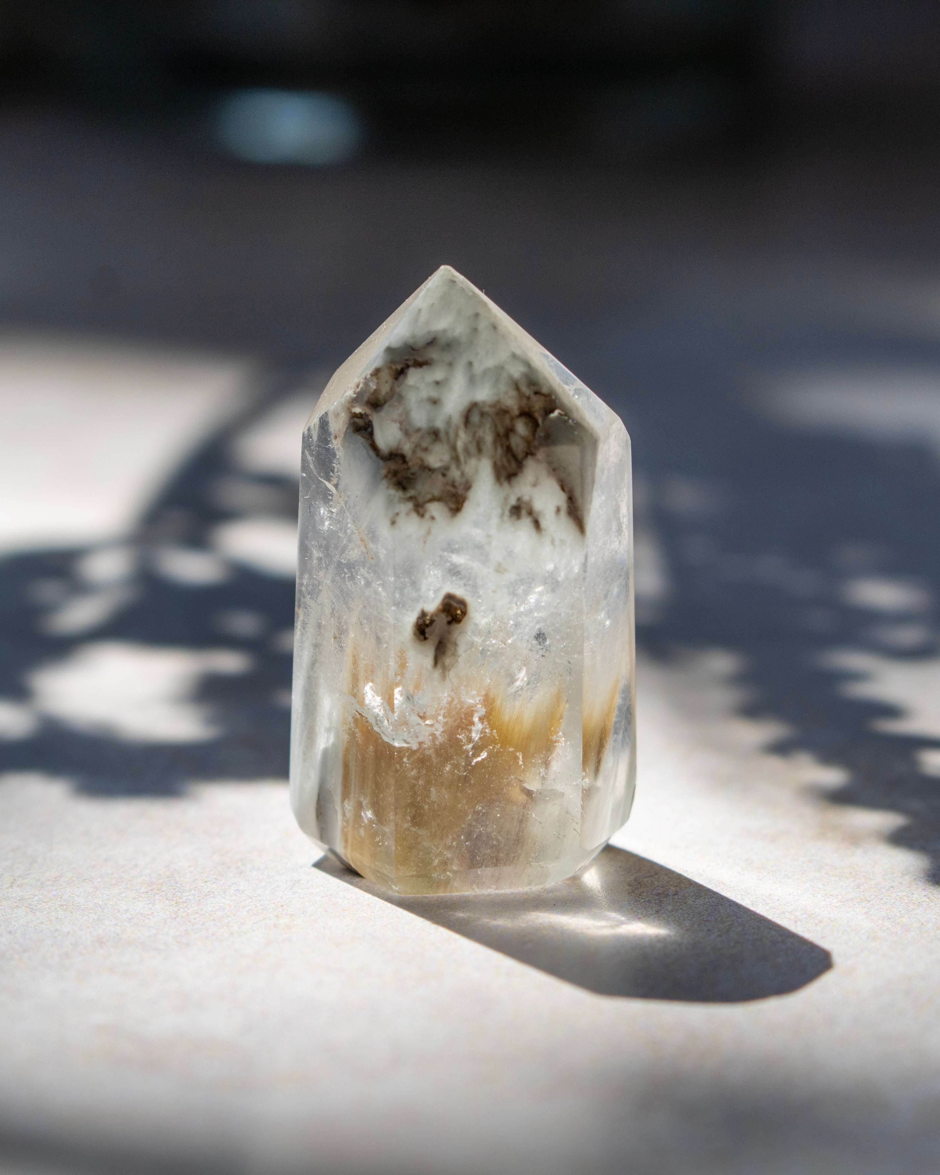 A natural clear quartz crystal point with brown and tan inclusions, standing upright on a light surface in direct sunlight with a sharp shadow.
