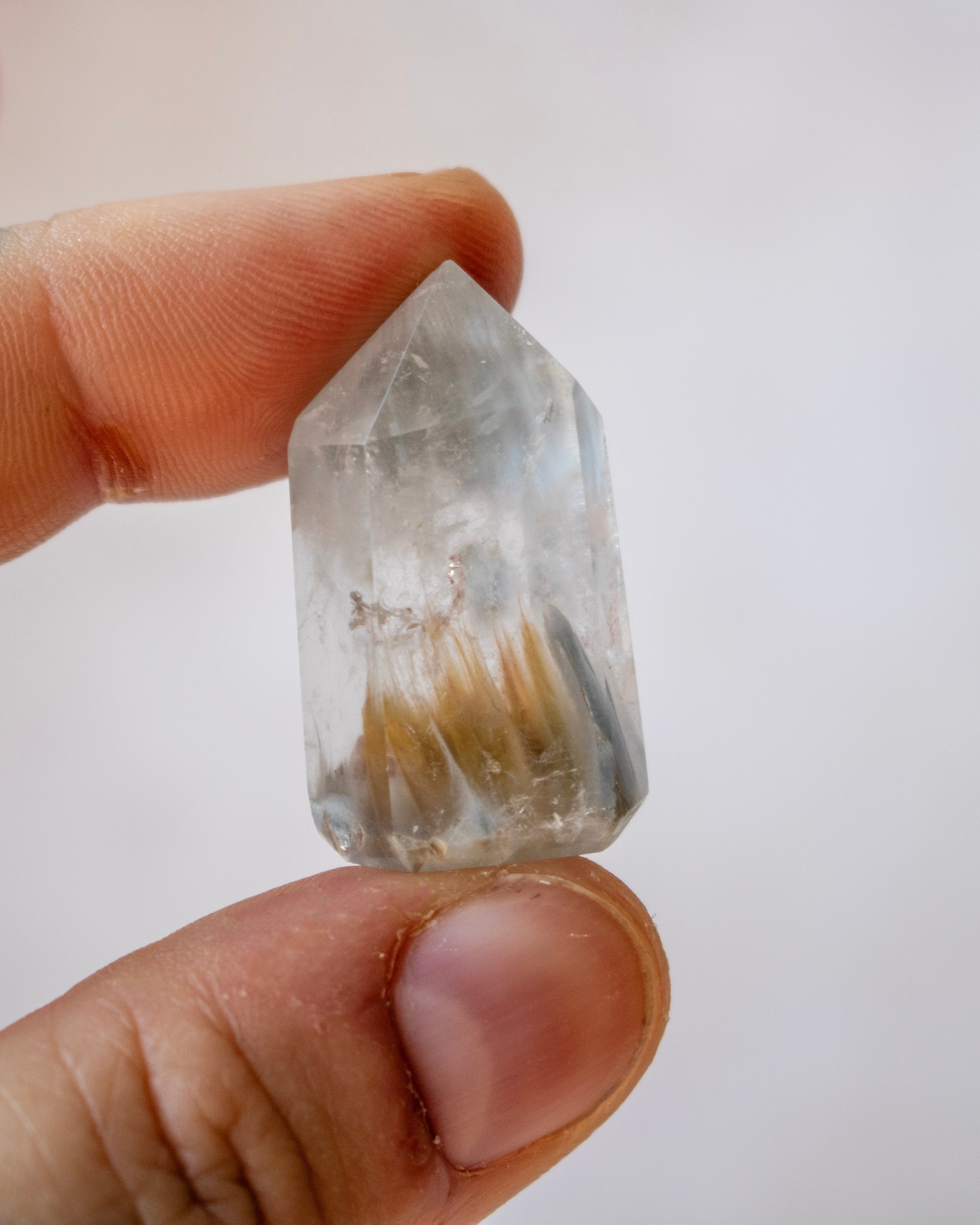A hand holding a clear pointed crystal with internal golden-brown and gray inclusions against a plain light background.