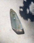 A natural translucent crystal point, likely agate, resting on a light beige textured surface with a prominent dark shadow cast across the right side.