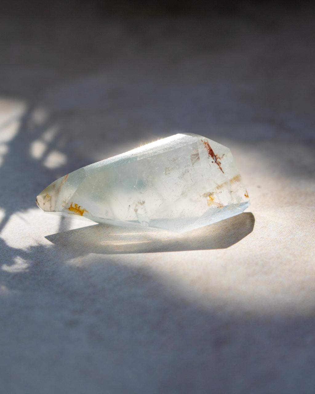 A raw, unpolished translucent crystal with natural inclusions and tiny brownish inclusions, resting on a textured gray surface under direct sunlight with a soft shadow.