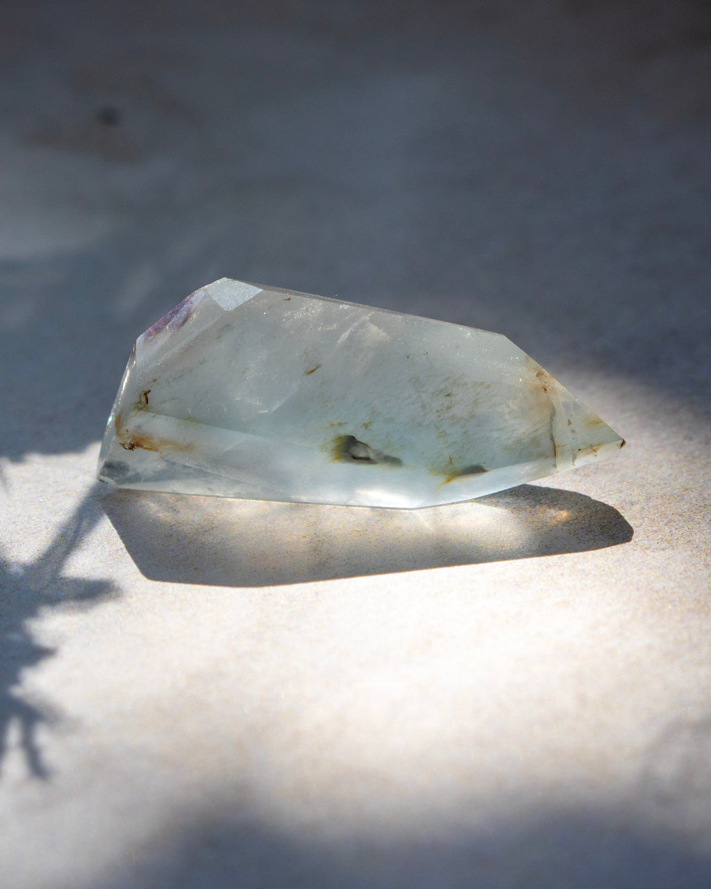 A natural clear quartz crystal point with inclusions, resting on a light surface under direct sunlight with a sharp shadow.