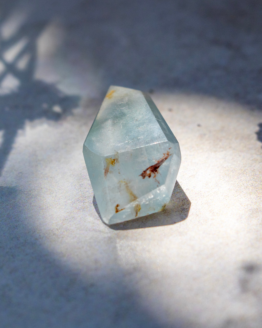 A faceted light blue-green crystal with natural inclusions and brown flecks, photographed on a textured light surface under direct sunlight with soft shadows.