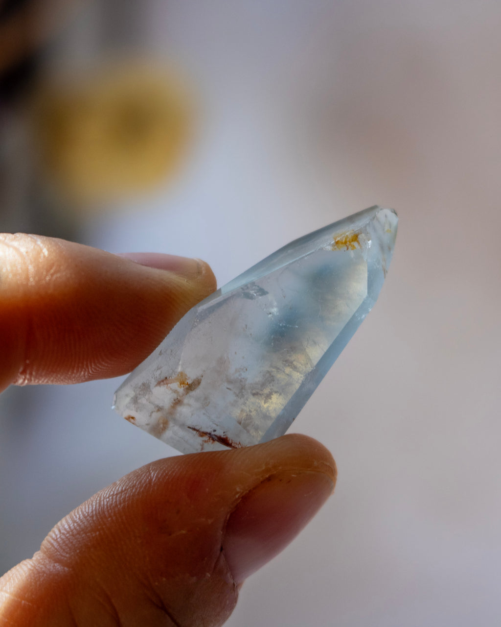 A hand holding a small, pointed, pale blue translucent crystal or gemstone against a softly blurred light background.