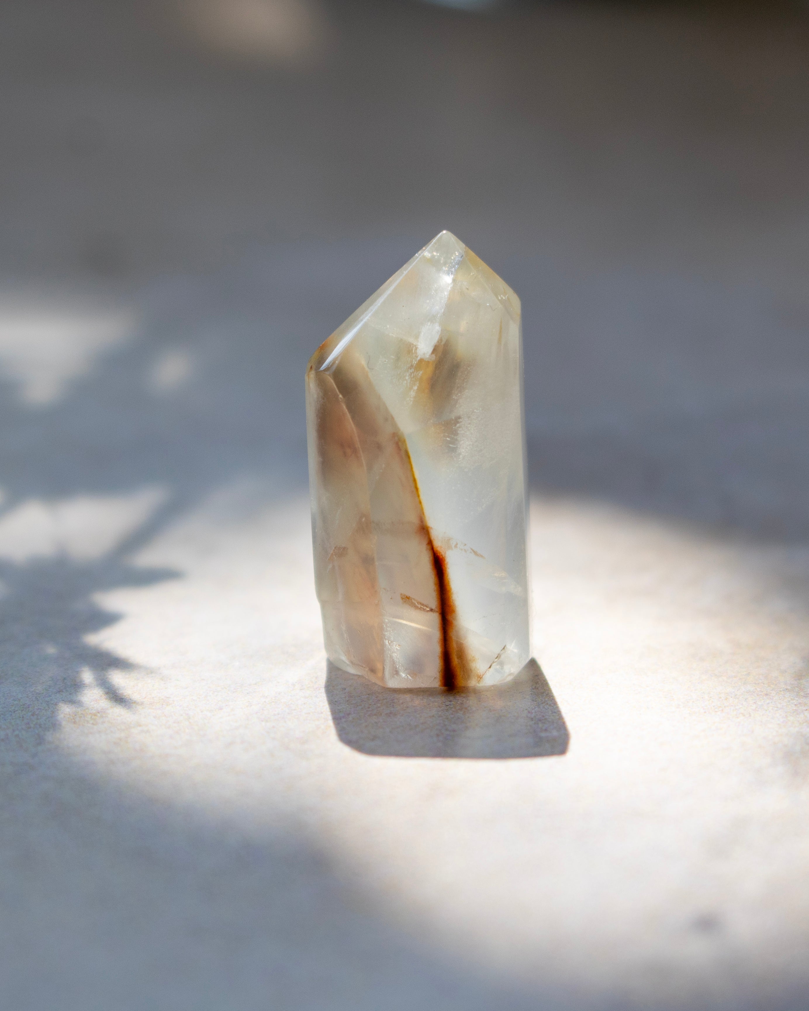 A natural pointed crystal stone with translucent milky white and warm brown inclusions, standing upright on a light surface under direct sunlight with a soft shadow.
