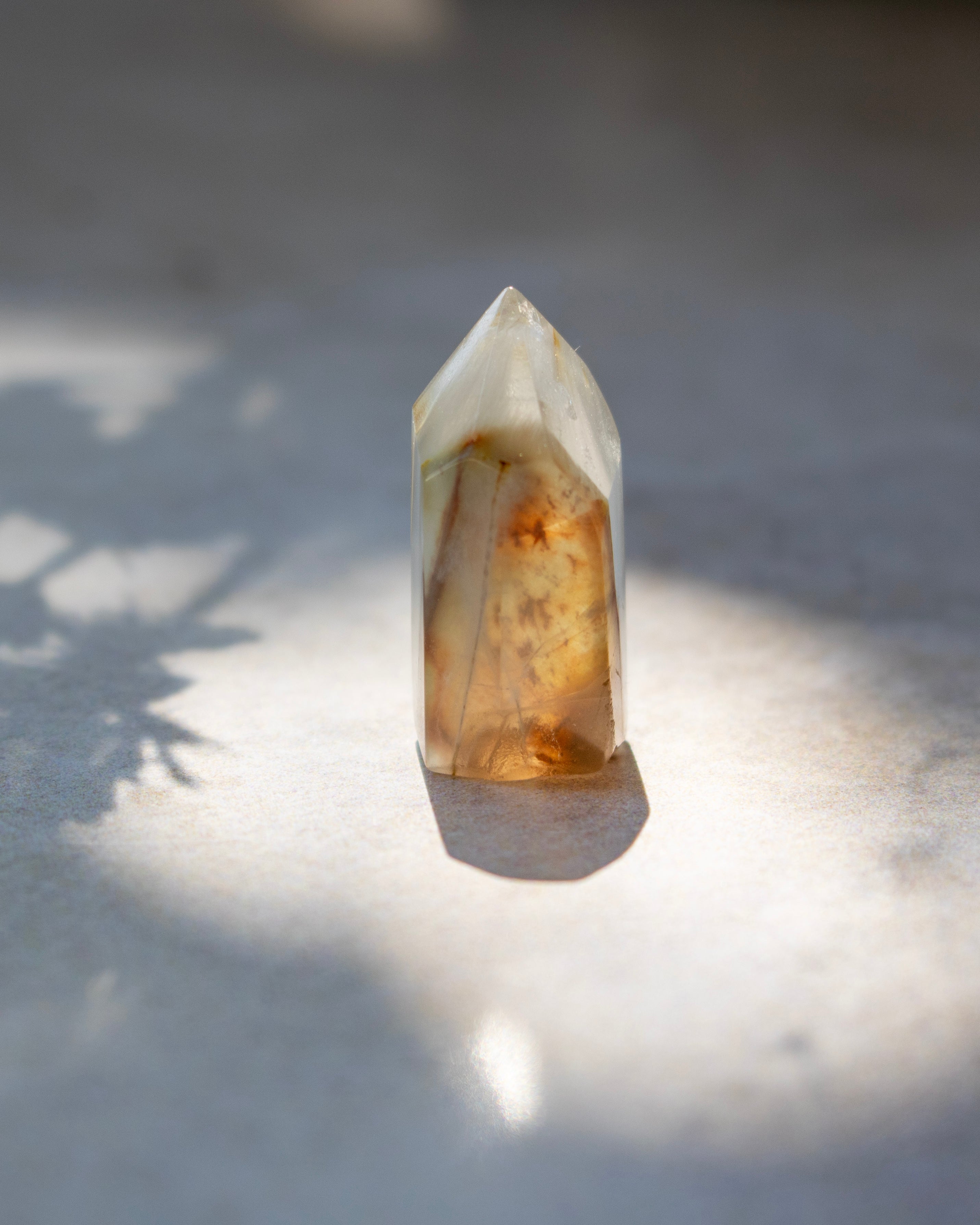 A polished pointed crystal tower with natural amber and brown inclusions, standing upright on a light surface under direct sunlight with soft shadows.