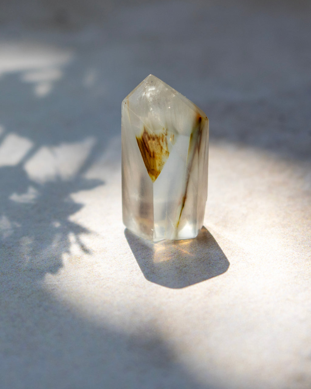 A clear quartz crystal point with brown and yellow inclusions, standing on a light textured surface under direct sunlight with leaf shadows.