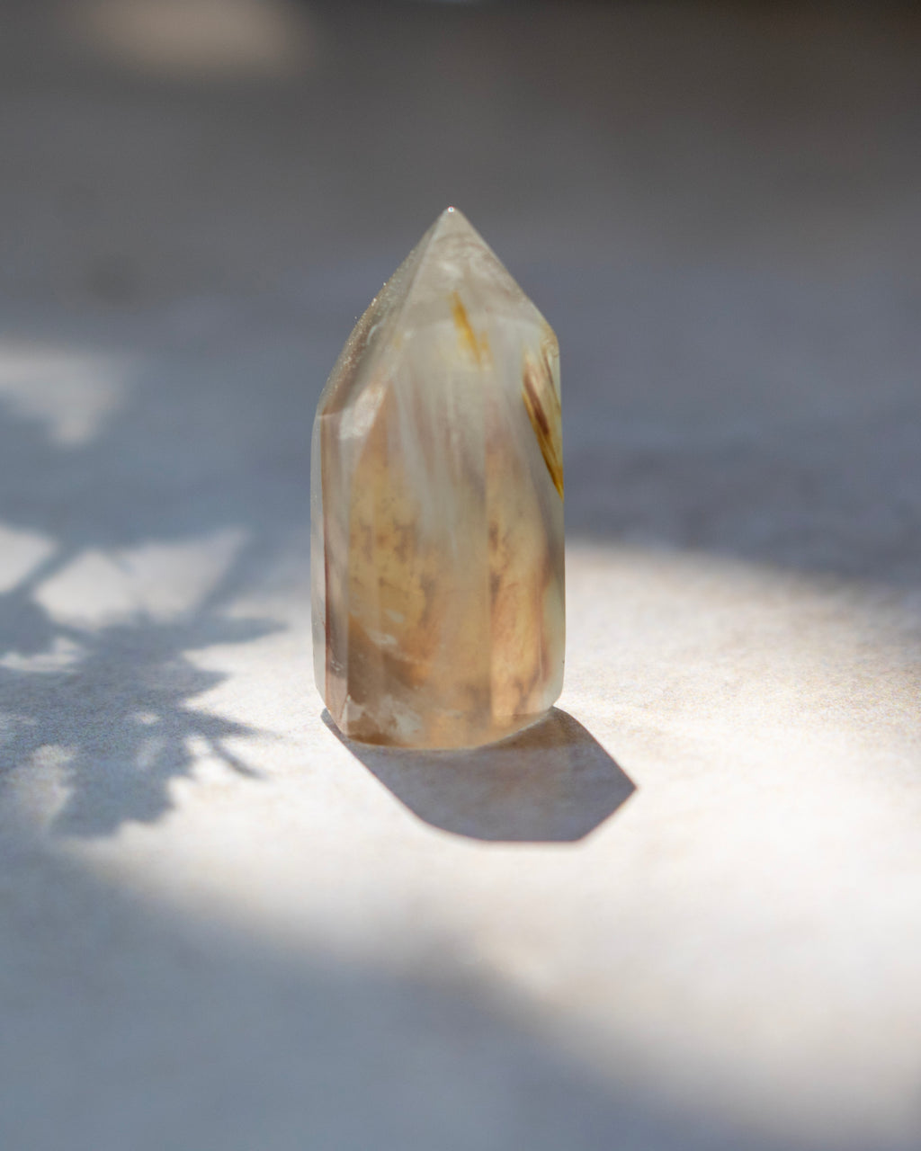 A natural pointed crystal tower with internal inclusions, standing on a light surface under direct sunlight that casts a shadow.